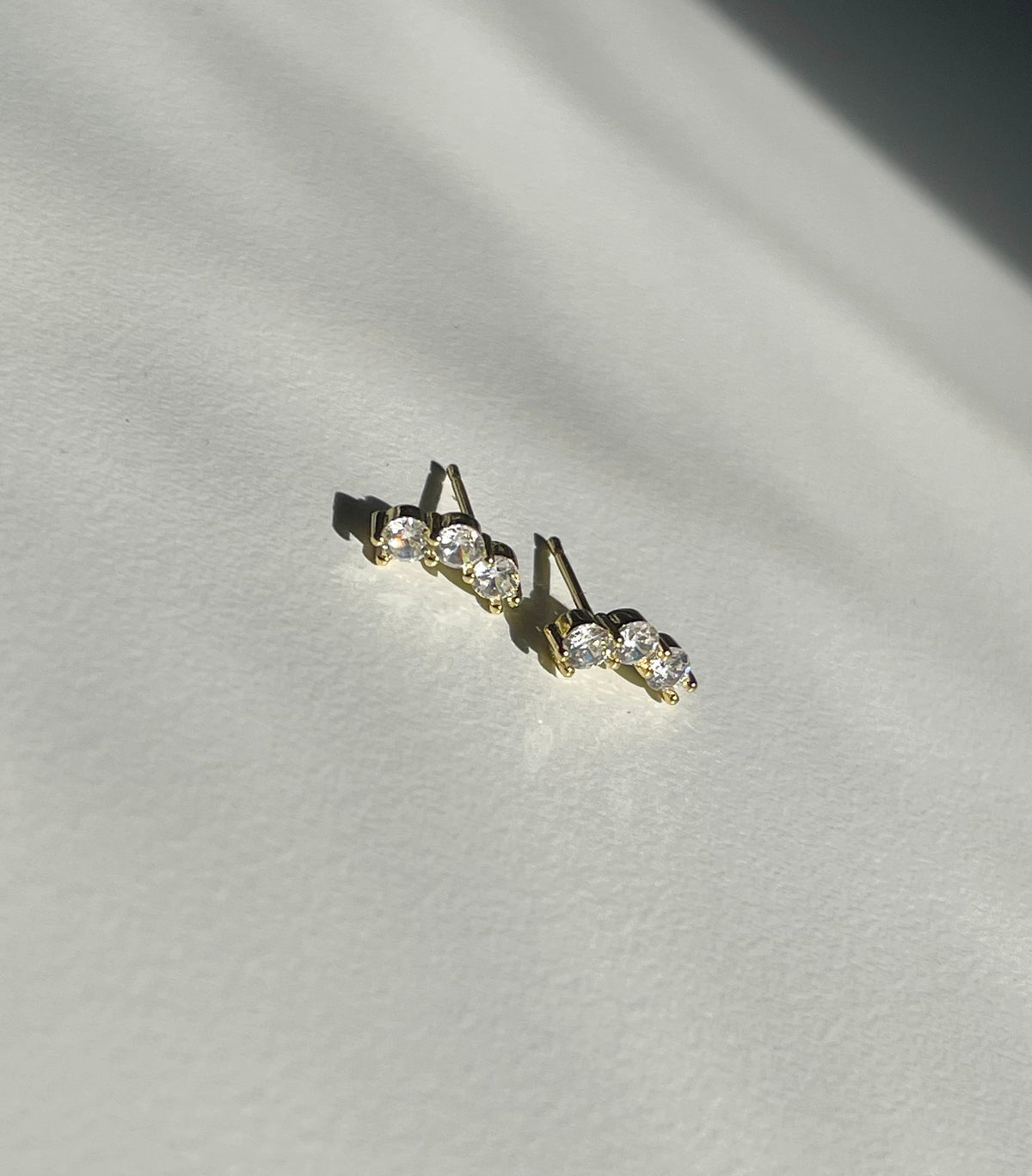 A pair of gold plated stud earrings with three clear round-cut stones each, displayed on a textured off-white surface with dramatic diagonal shadows.