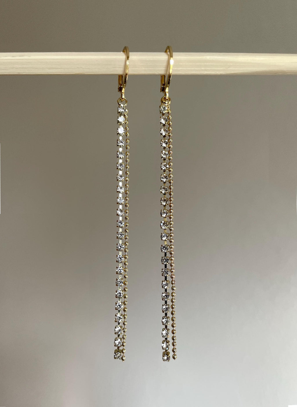 A pair of gold-tone drop earrings with multiple rows of clear rhinestones, hanging from a light-colored wooden dowel against a neutral gray background.