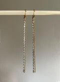 A pair of gold-tone drop earrings with multiple rows of clear rhinestones, hanging from a light-colored wooden dowel against a neutral gray background.