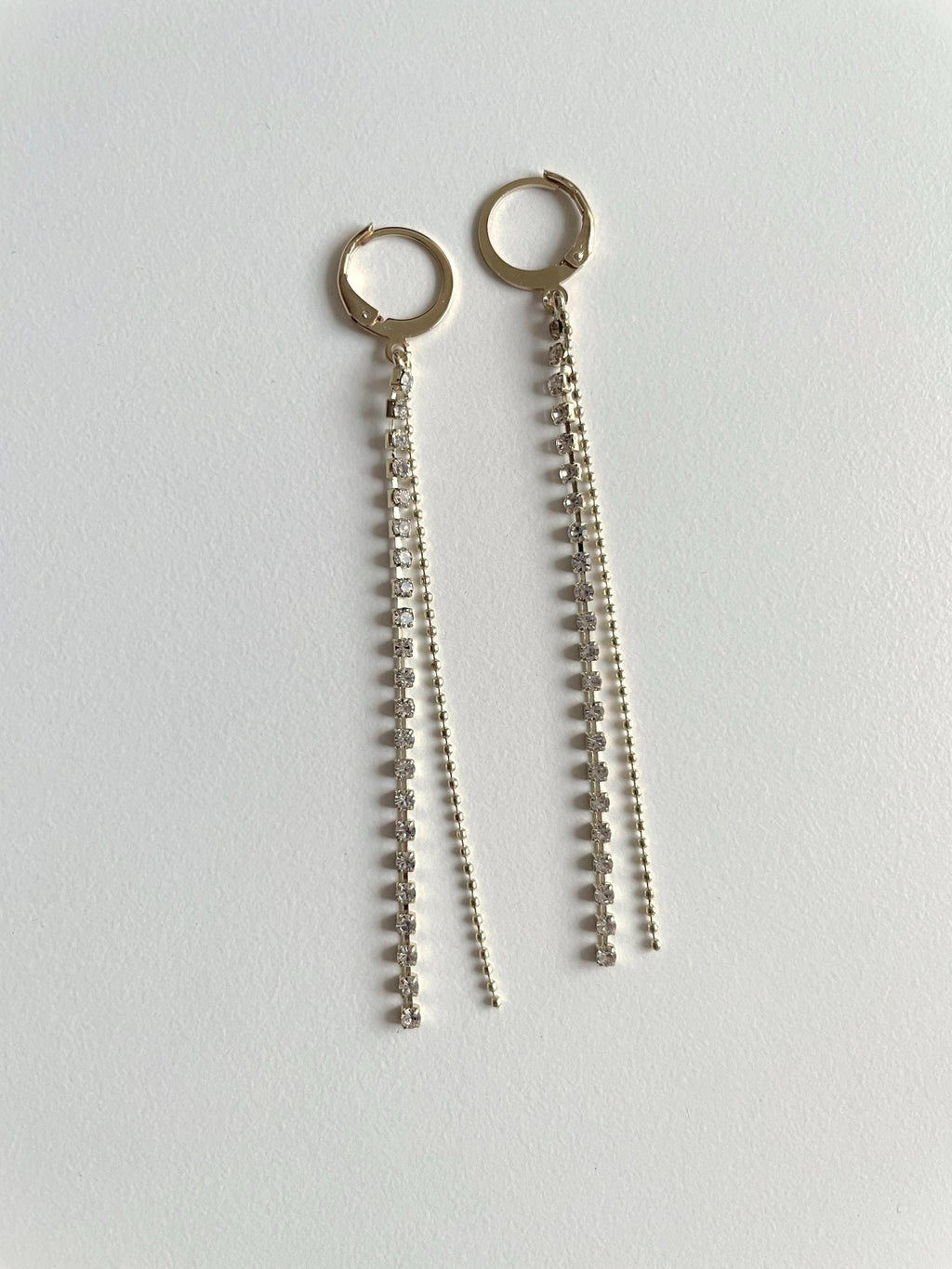 A pair of gold-toned huggie hoop earrings with long dangling chain and bead fringe, displayed on a plain light gray surface.
