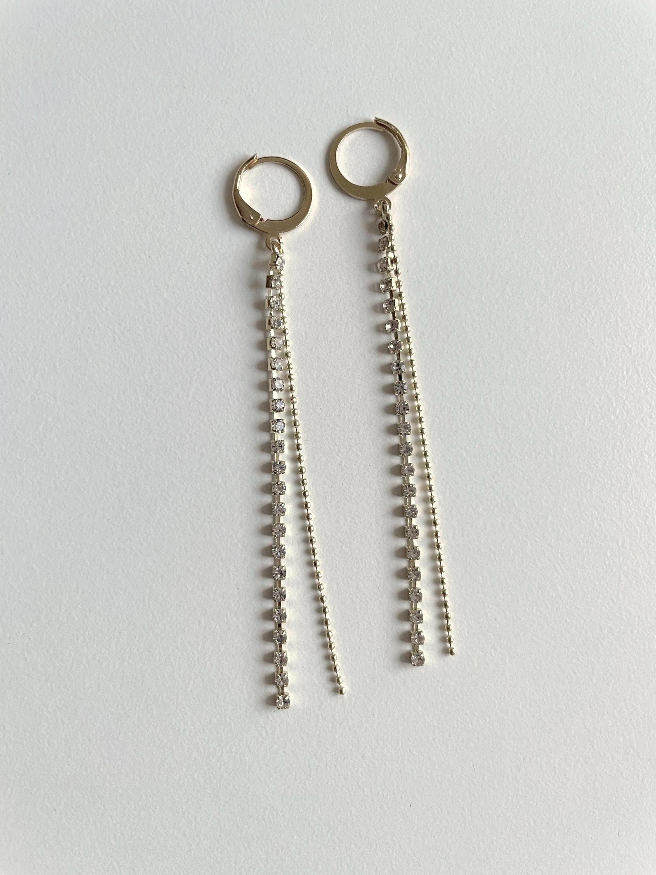 A pair of gold-toned huggie hoop earrings with long dangling chain and bead fringe, displayed on a plain light gray surface.