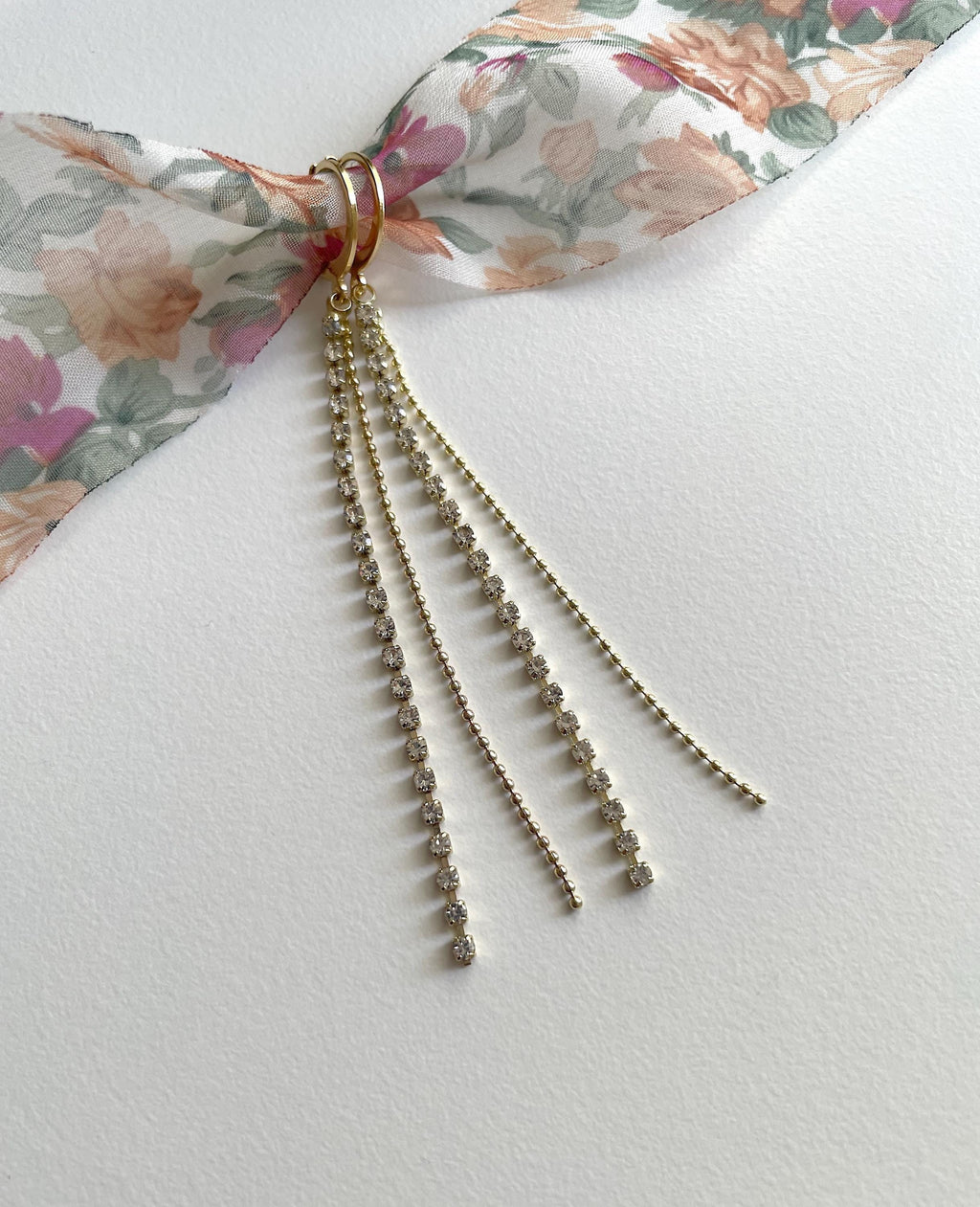 A gold-toned ring with three dangling chain strands, each studded with small clear beads, tied to a floral ribbon on a white textured background.