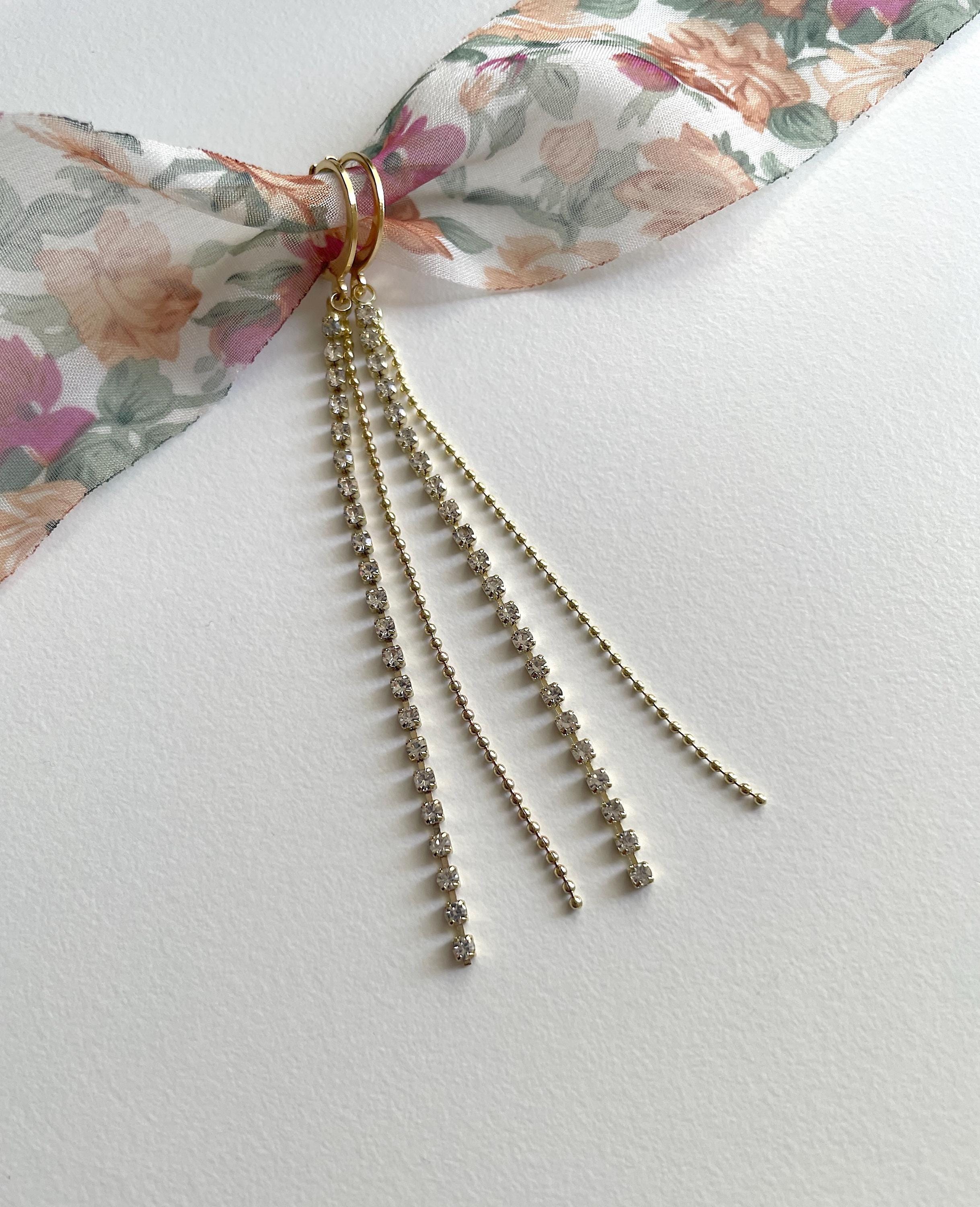 A gold-toned ring with three dangling chain strands, each studded with small clear beads, tied to a floral ribbon on a white textured background.