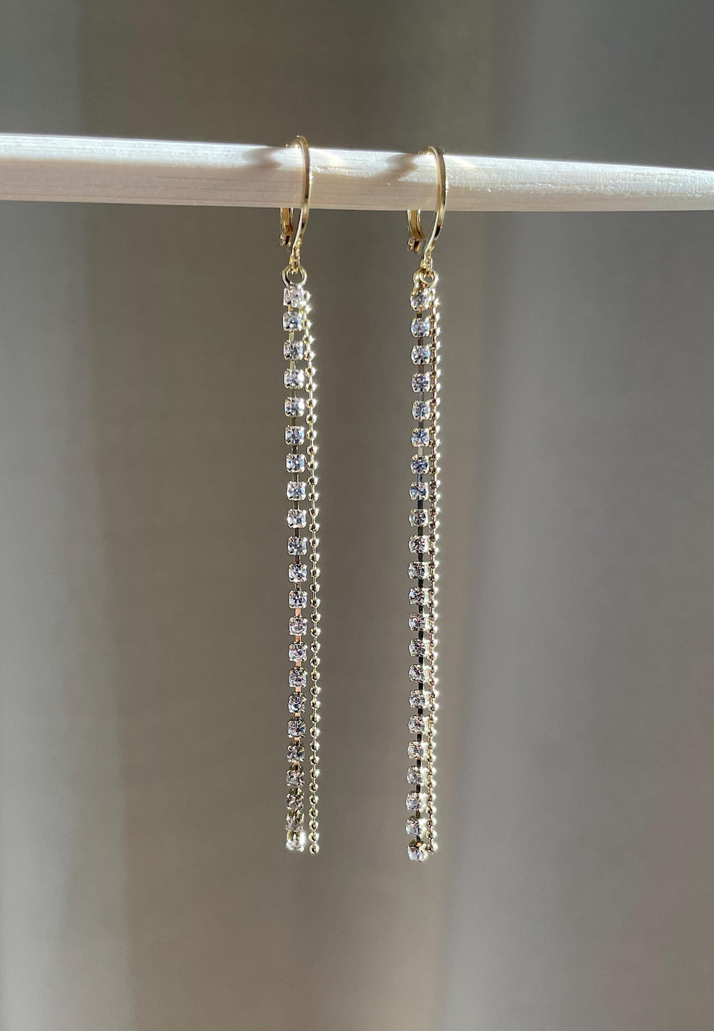A pair of gold-tone dangle earrings with clear rhinestones, hanging from a white display stand against a soft gray background.