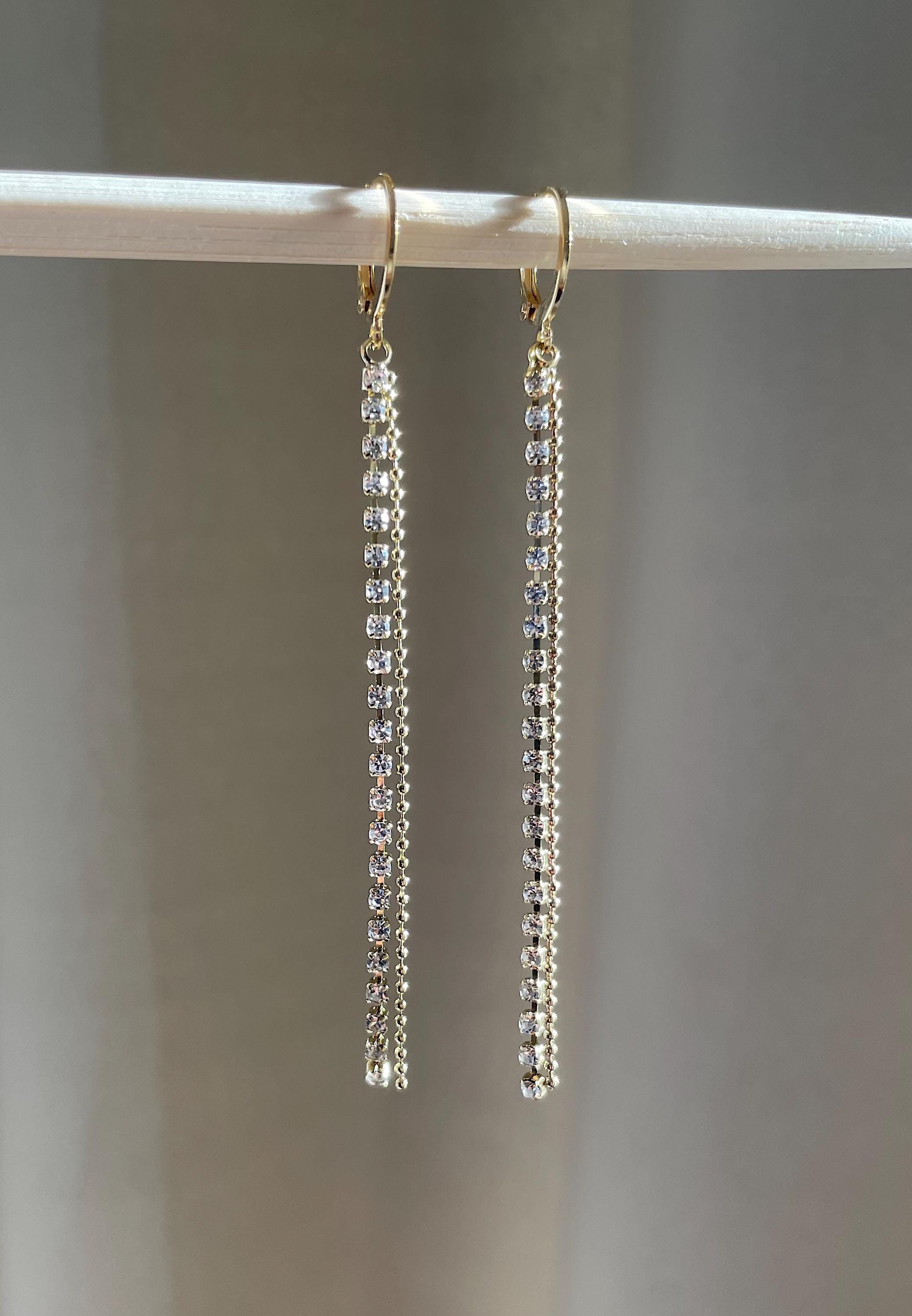 A pair of gold-tone dangle earrings with clear rhinestones, hanging from a white display stand against a soft gray background.