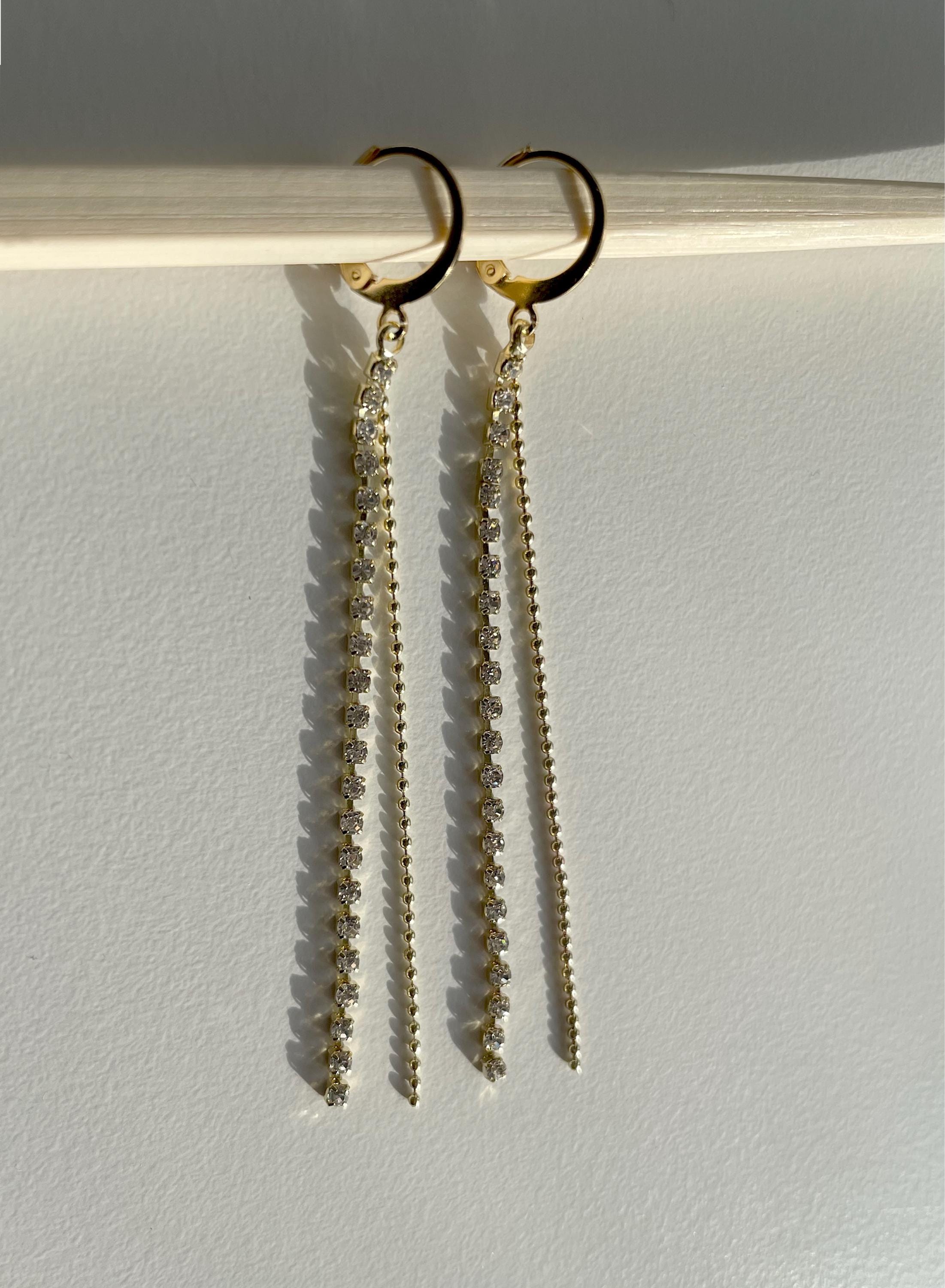 A pair of gold huggie hoop earrings with long, beaded drop chains, hanging from a light-colored wooden rod against a plain white wall.