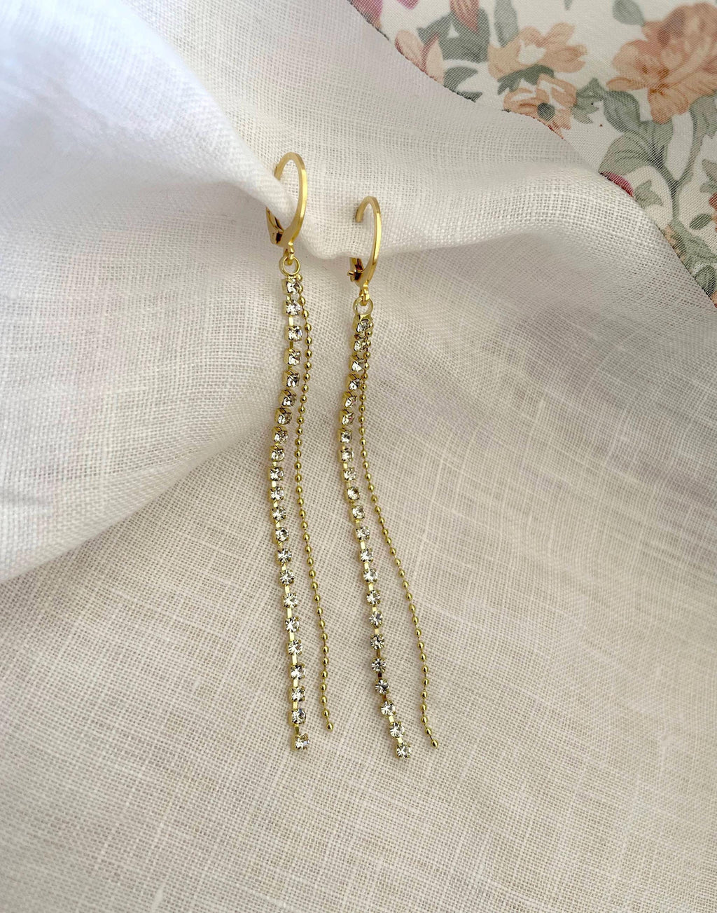 A pair of gold-toned dangle earrings with small clear stones, displayed on draped white fabric with a floral print visible in the top right corner.