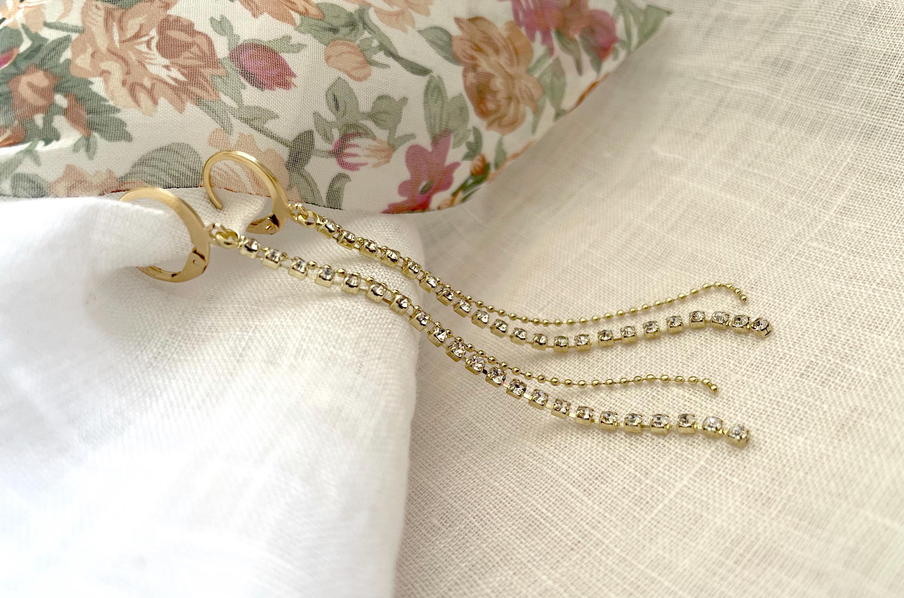 Gold-toned dangle earrings with small clear rhinestone accents, displayed on a textured cream fabric with a floral fabric prop in the background.