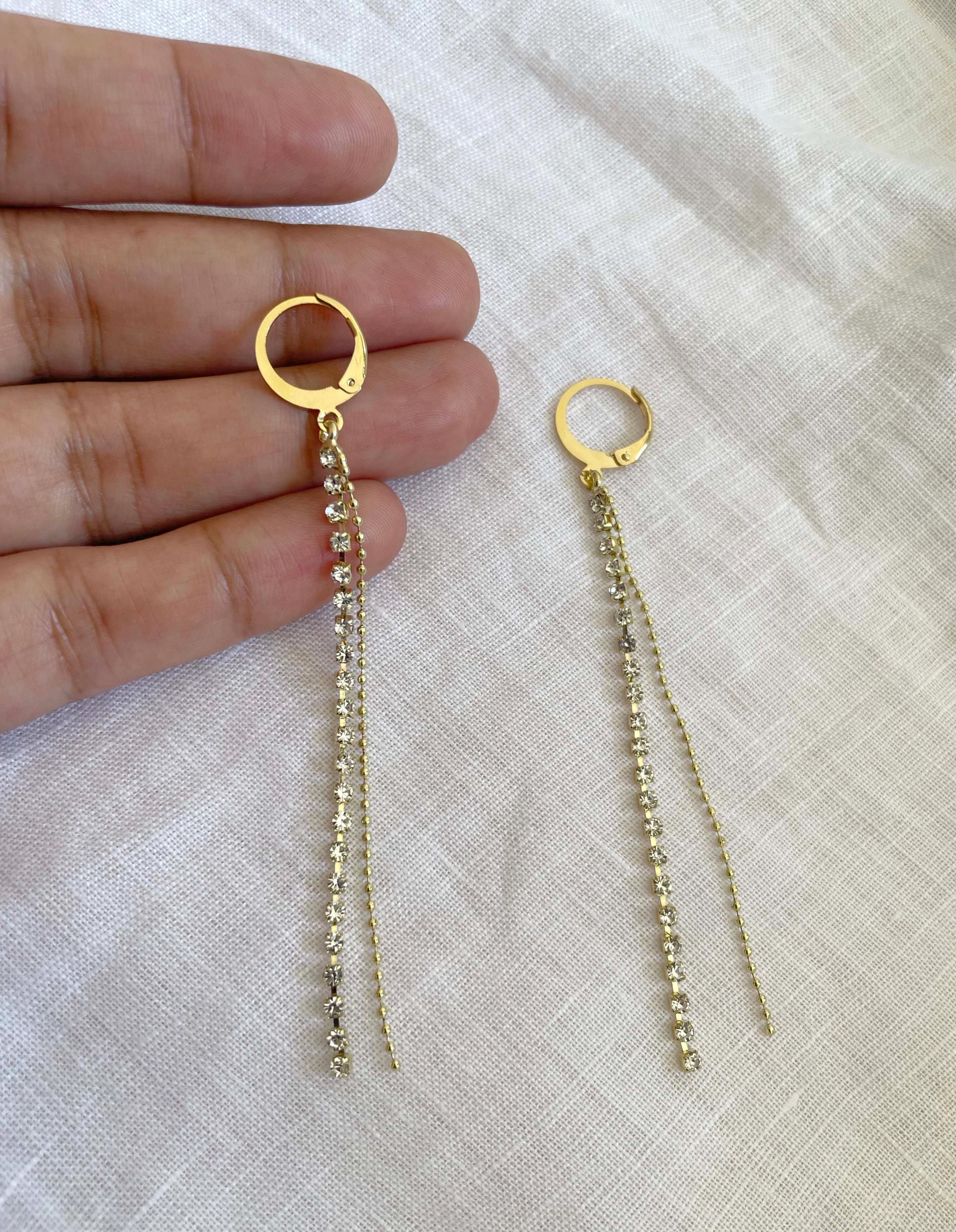 A pair of gold hoop dangle earrings with small clear stones and long chain drops, shown with a person's hand holding one earring against a light fabric background.