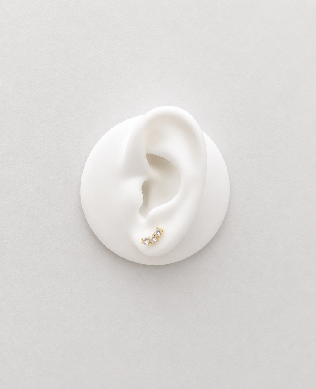A small gold plated crystal studs with tiny clear stones, displayed on a white ear-shaped mannequin against a light gray background.