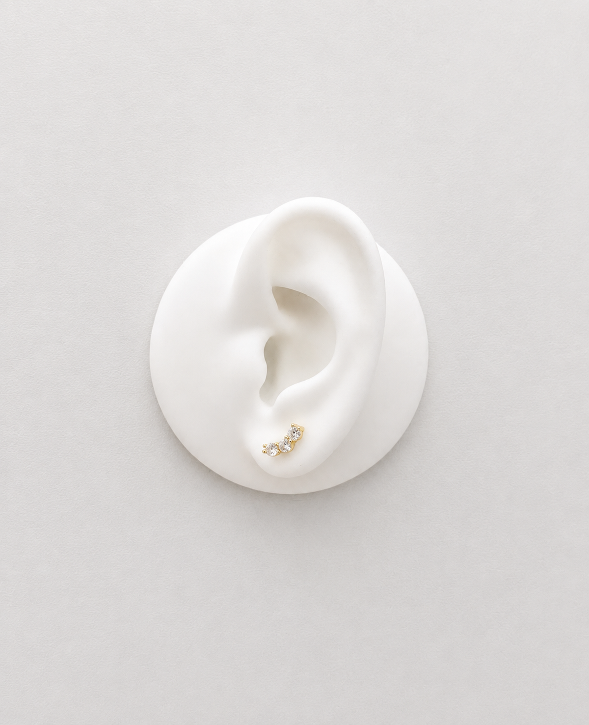 A small gold plated crystal studs with tiny clear stones, displayed on a white ear-shaped mannequin against a light gray background.