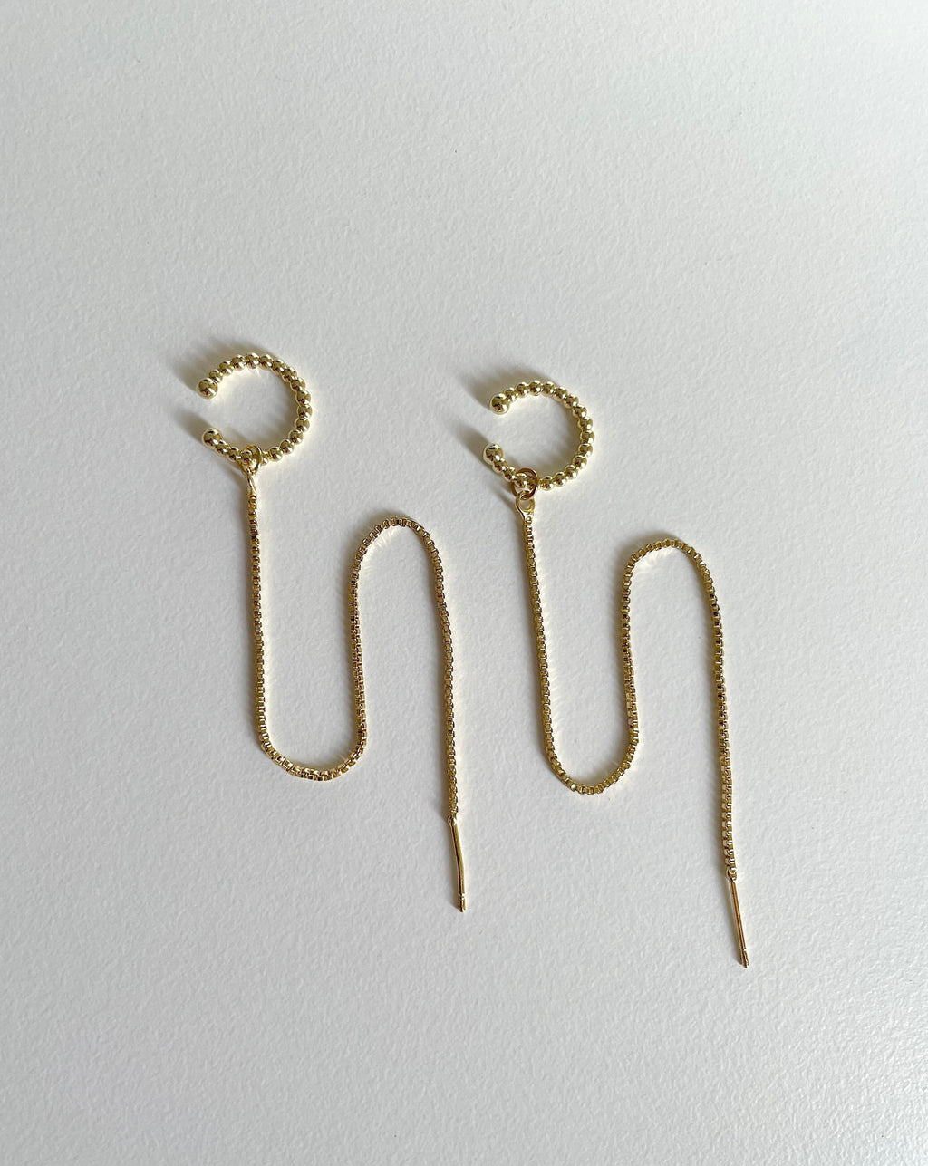 A pair of gold plated cuff chain earrings with textured circular cuffs and long wavy drop chains ending in pointed tips, displayed on a plain light gray surface.