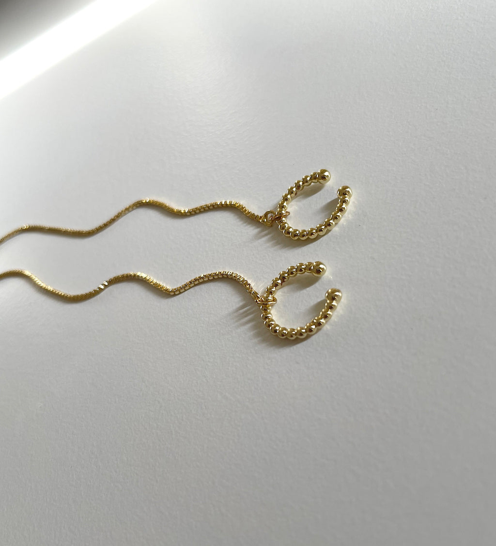 A pair of gold wavy cuff chain earrings with textured beaded ends, displayed on a clean white surface.