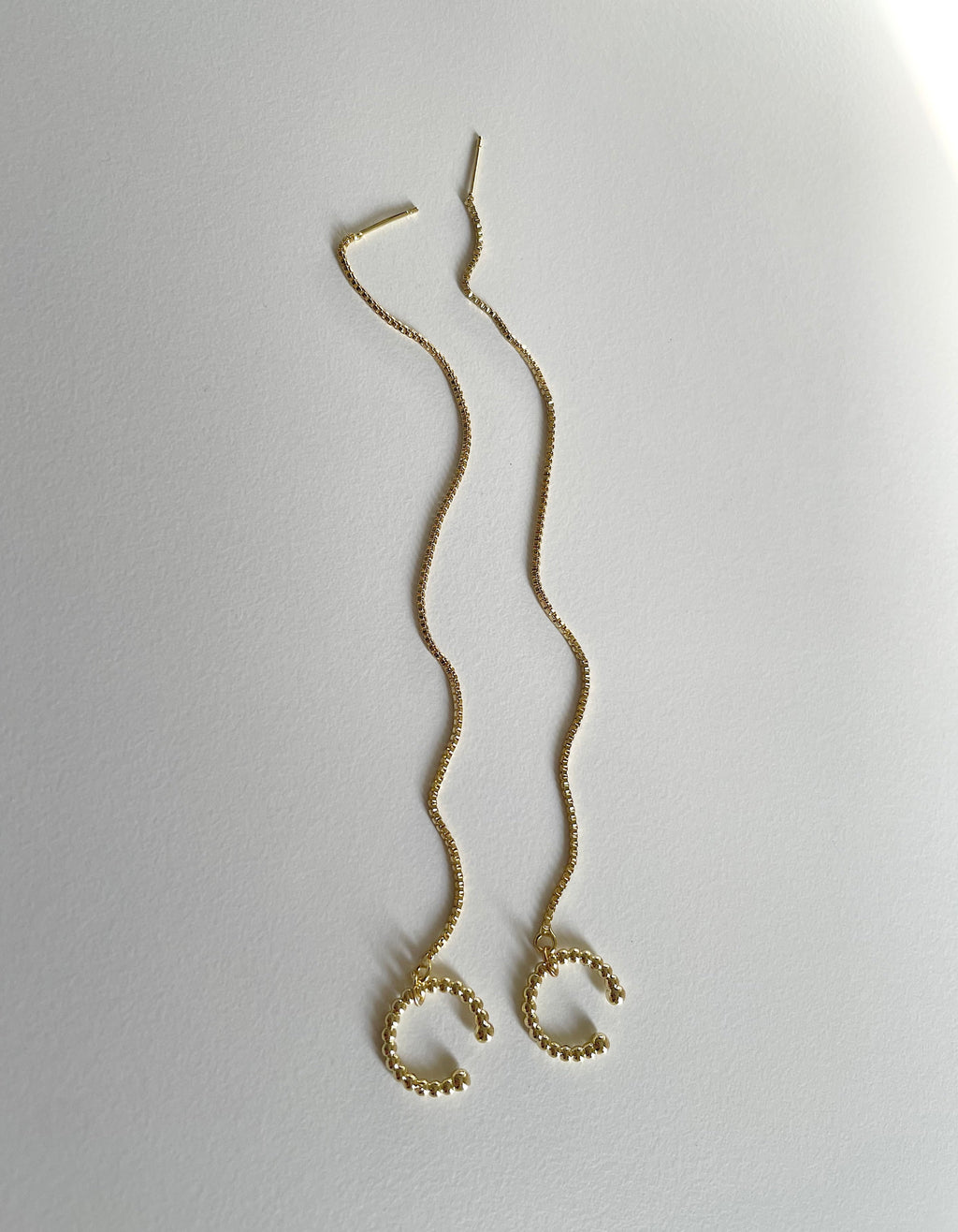 A pair of delicate gold wavy threader earrings with small beaded circular cuff, displayed on a light gray textured background.