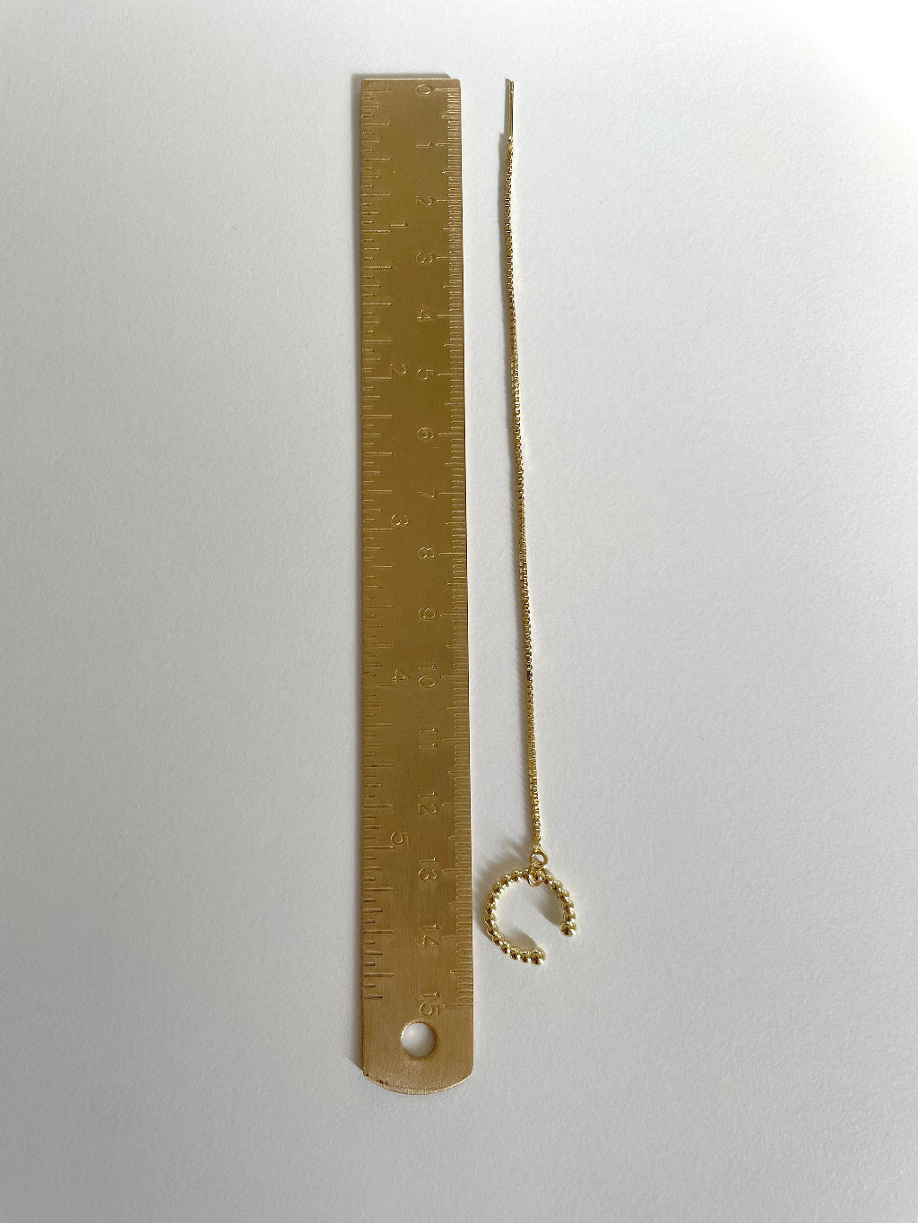 A gold plated cuff earring with a small textured cuff and long delicate threader chain, shown next to a golden metal ruler for scale on a plain white background.