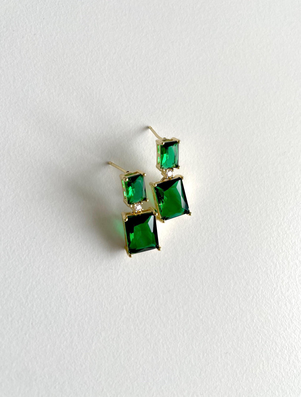 A pair of gold-tone drop earrings with rectangular green gemstones, displayed on a textured white background.