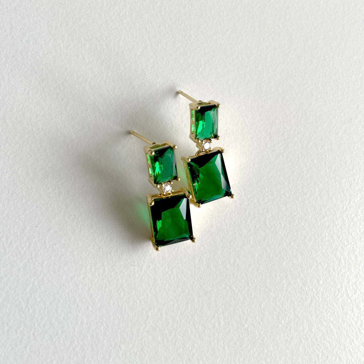 A pair of gold plated drop earrings featuring rectangular green emerald gemstones in a stacked design, displayed on a textured white surface.