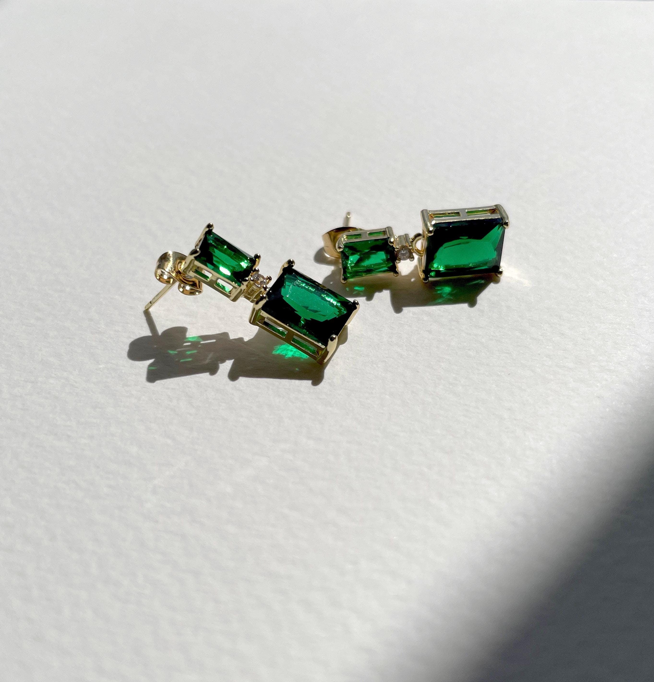 A pair of gold-tone stud earrings with rectangular green gemstones, displayed on a textured white surface with a soft shadow.