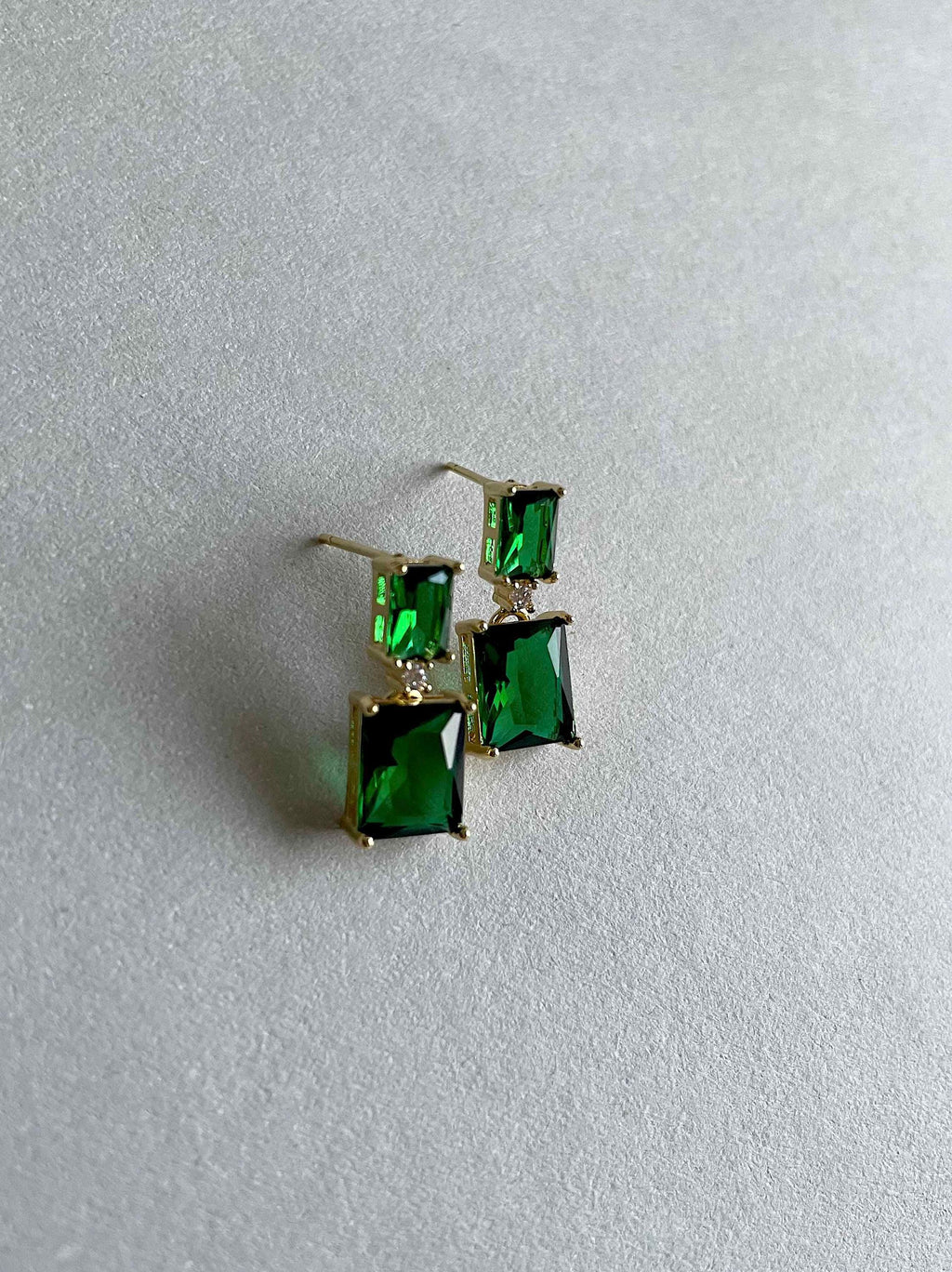 A pair of gold-tone drop earrings with rectangular green gemstones, resting on a textured light gray surface.