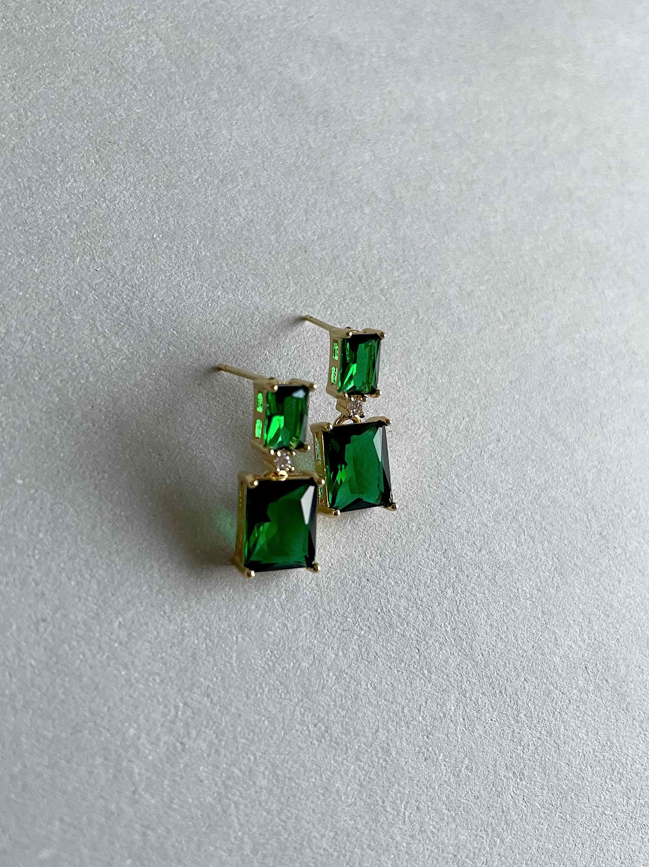 A pair of gold-tone drop earrings with rectangular green gemstones, resting on a textured light gray surface.