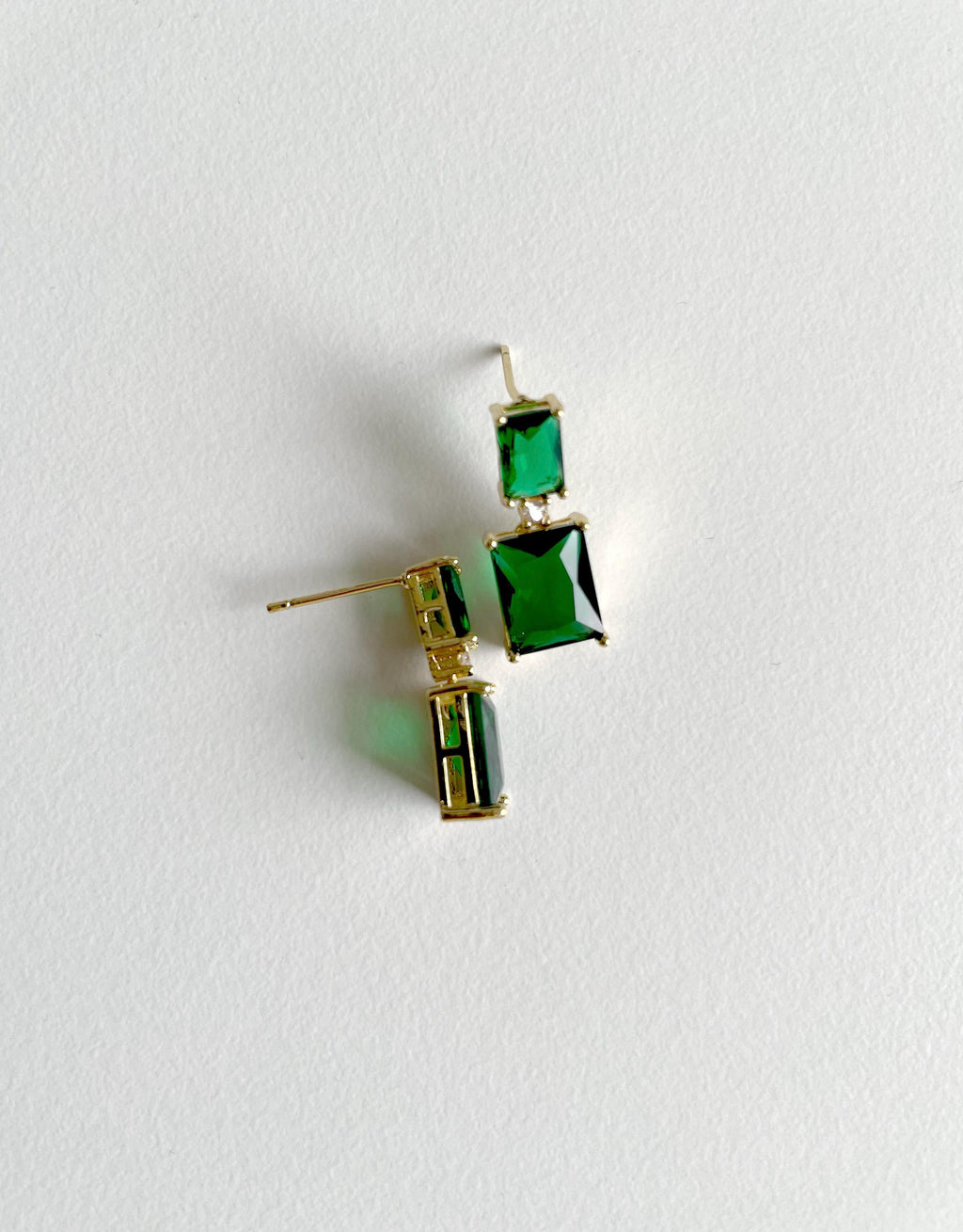 A pair of gold-tone drop earrings with rectangular green gemstones, displayed on a textured white background.