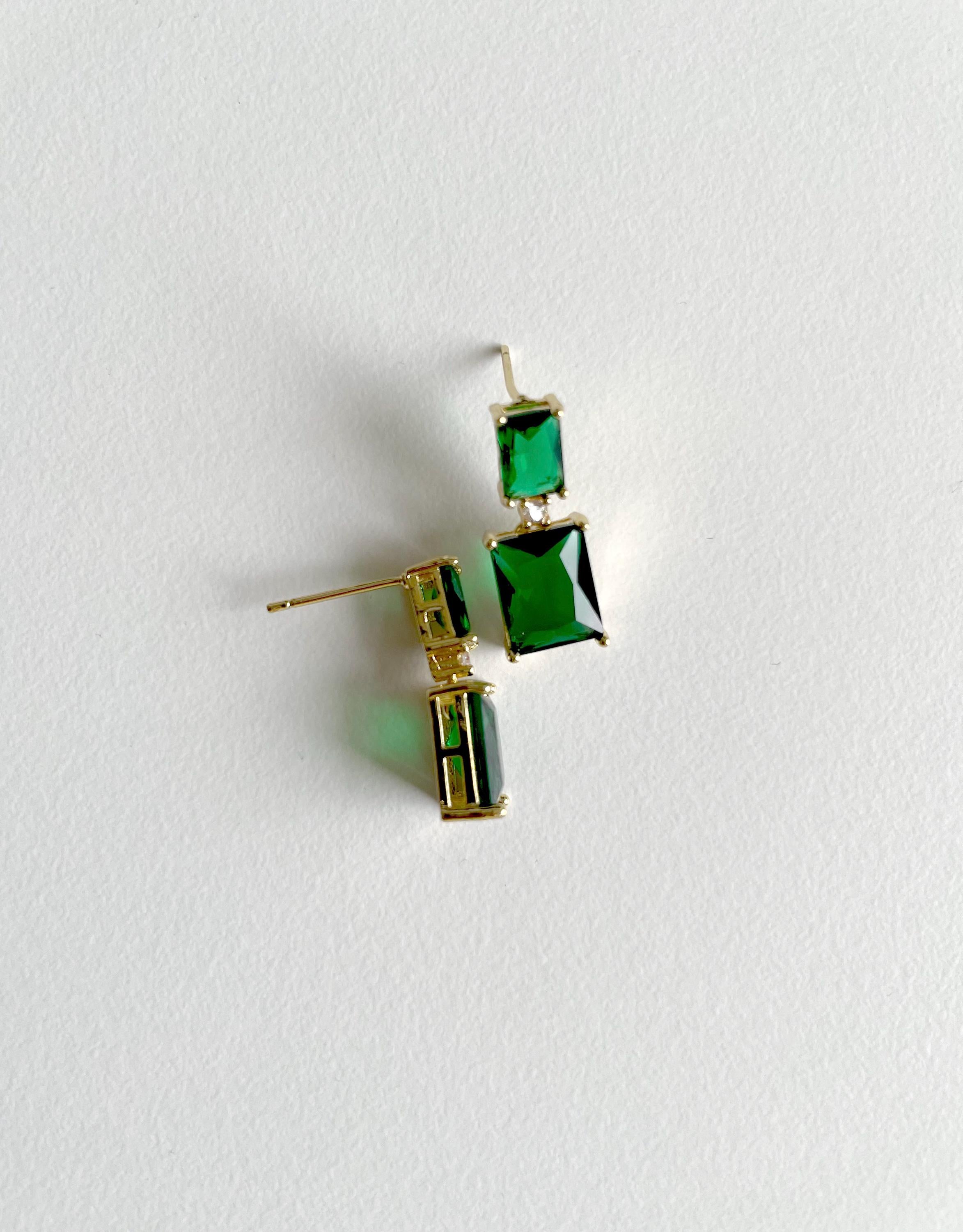 A pair of gold-tone drop earrings with rectangular green gemstones, displayed on a textured white background.