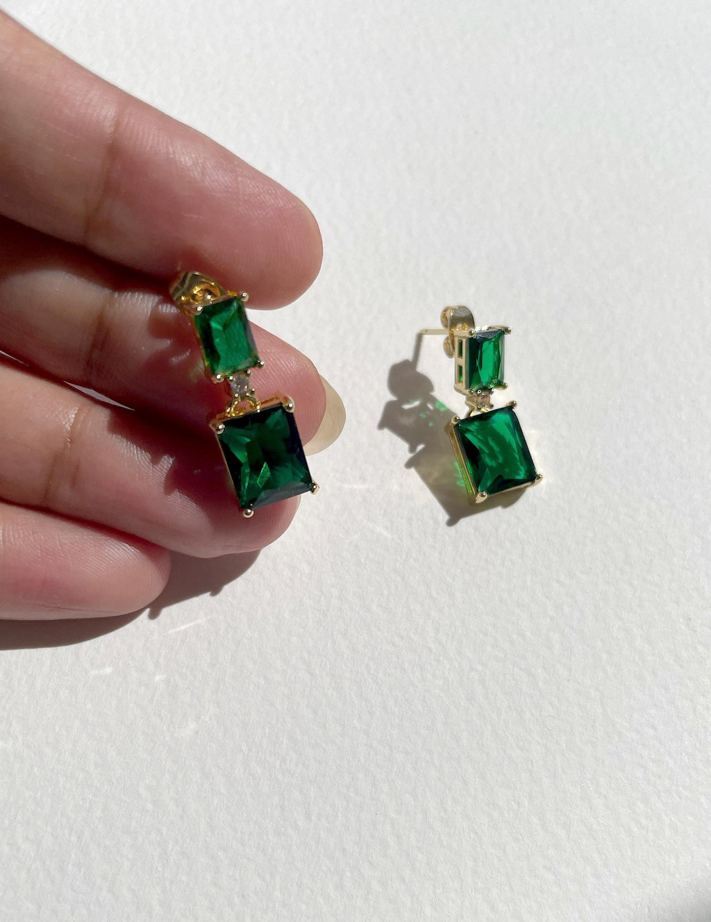 A pair of gold-plated drop earrings with green rectangular and square gemstones, shown with one earring held by a hand and the other resting on a white surface.