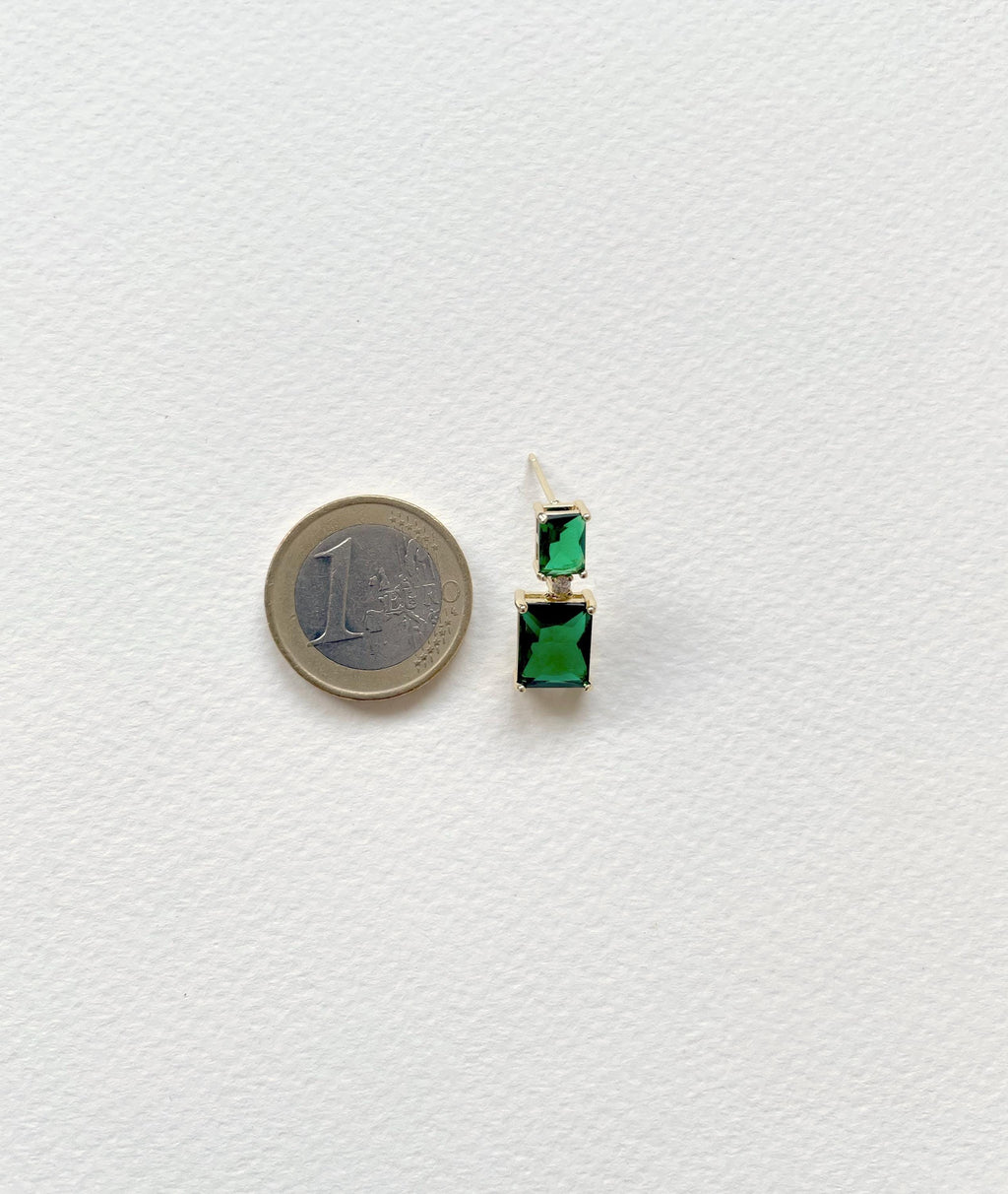 A single drop earring with a gold-toned post and two stacked rectangular green gemstones, shown next to a 1 Euro coin for scale on a textured white background.