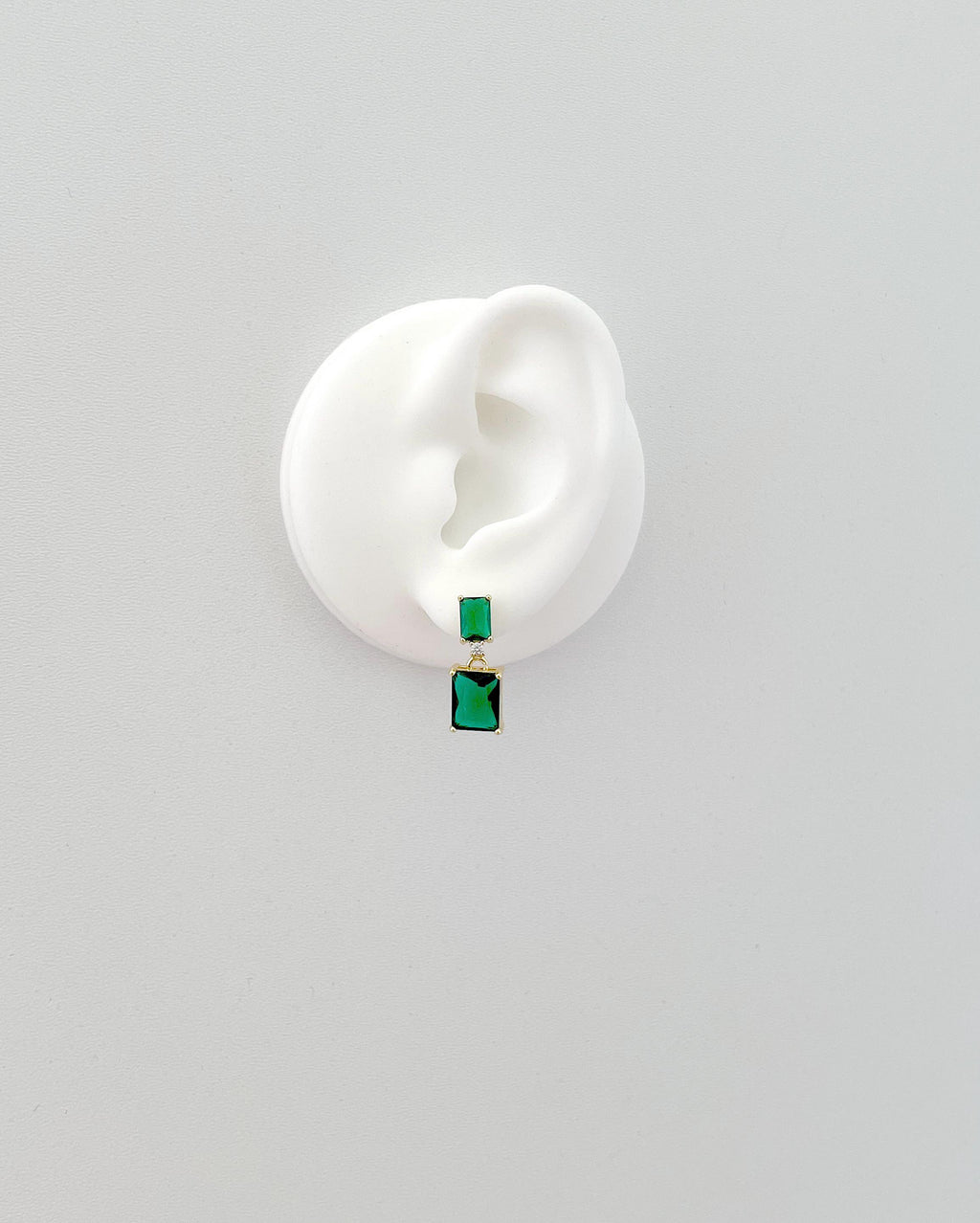 A single drop earring with a small green rectangular stone and a larger green marquise-cut drop, displayed on a white ear-shaped mannequin against a light gray background.