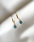 A pair of gold plated dangle earrings with circular blue and black evil eye charms, displayed on a textured white fabric background.