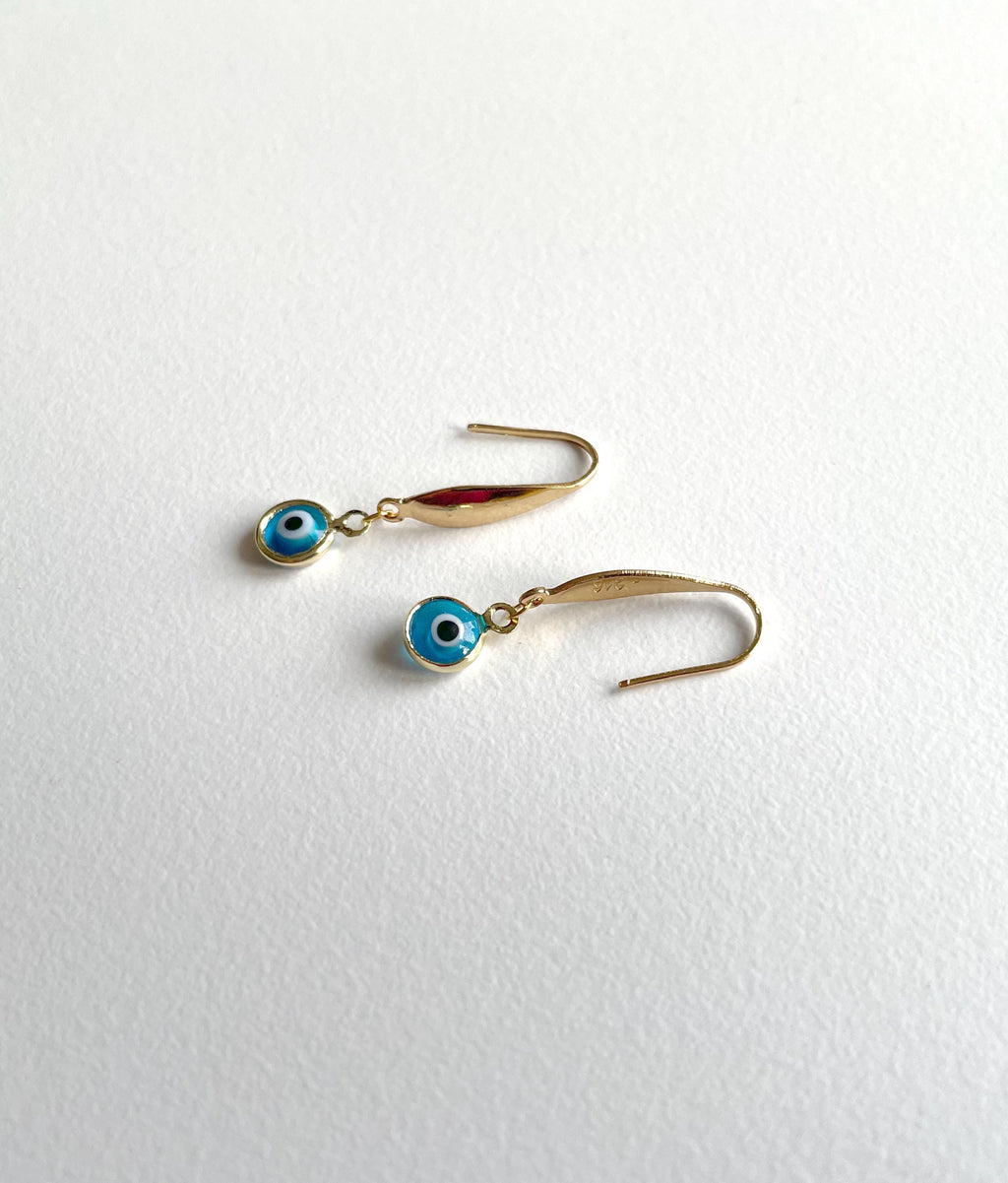A pair of gold-tone dangle earrings with small evil eye charms, displayed on a textured white surface.