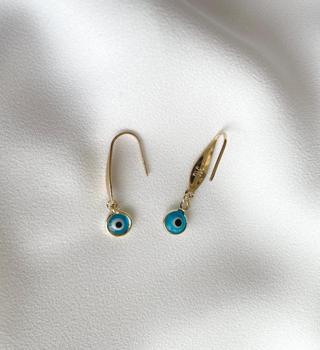 A pair of gold-tone dangle earrings with turquoise blue evil eye charms, displayed on a soft, white textured fabric background.