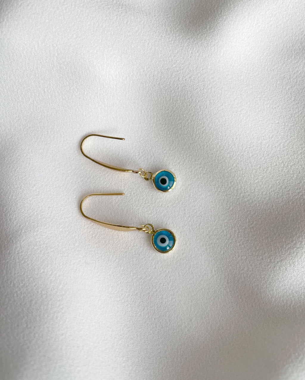A pair of gold-tone dangle earrings with small blue evil eye charms, displayed on a soft, textured white fabric background.