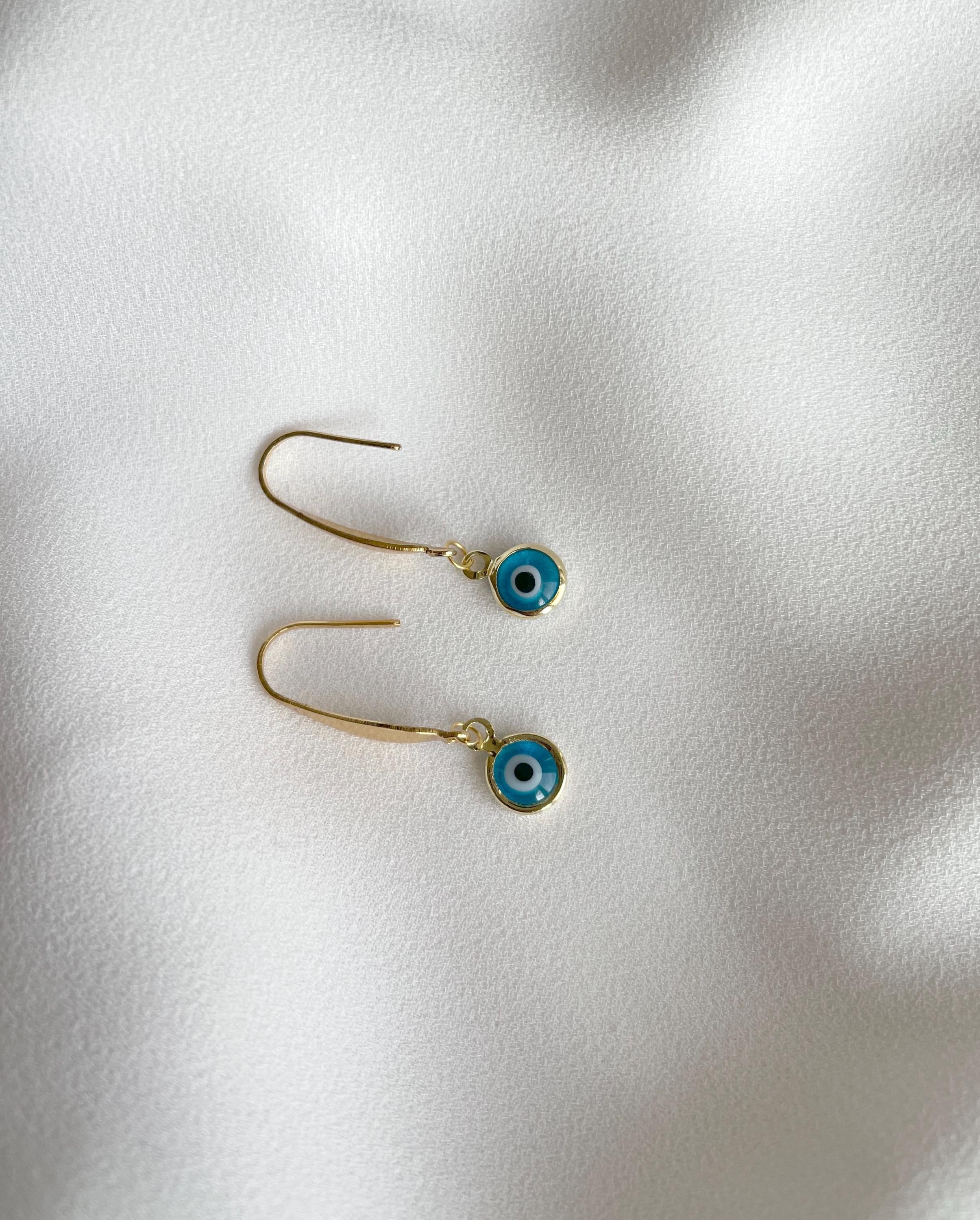 A pair of gold-tone dangle earrings with small blue evil eye charms, displayed on a soft, textured white fabric background.