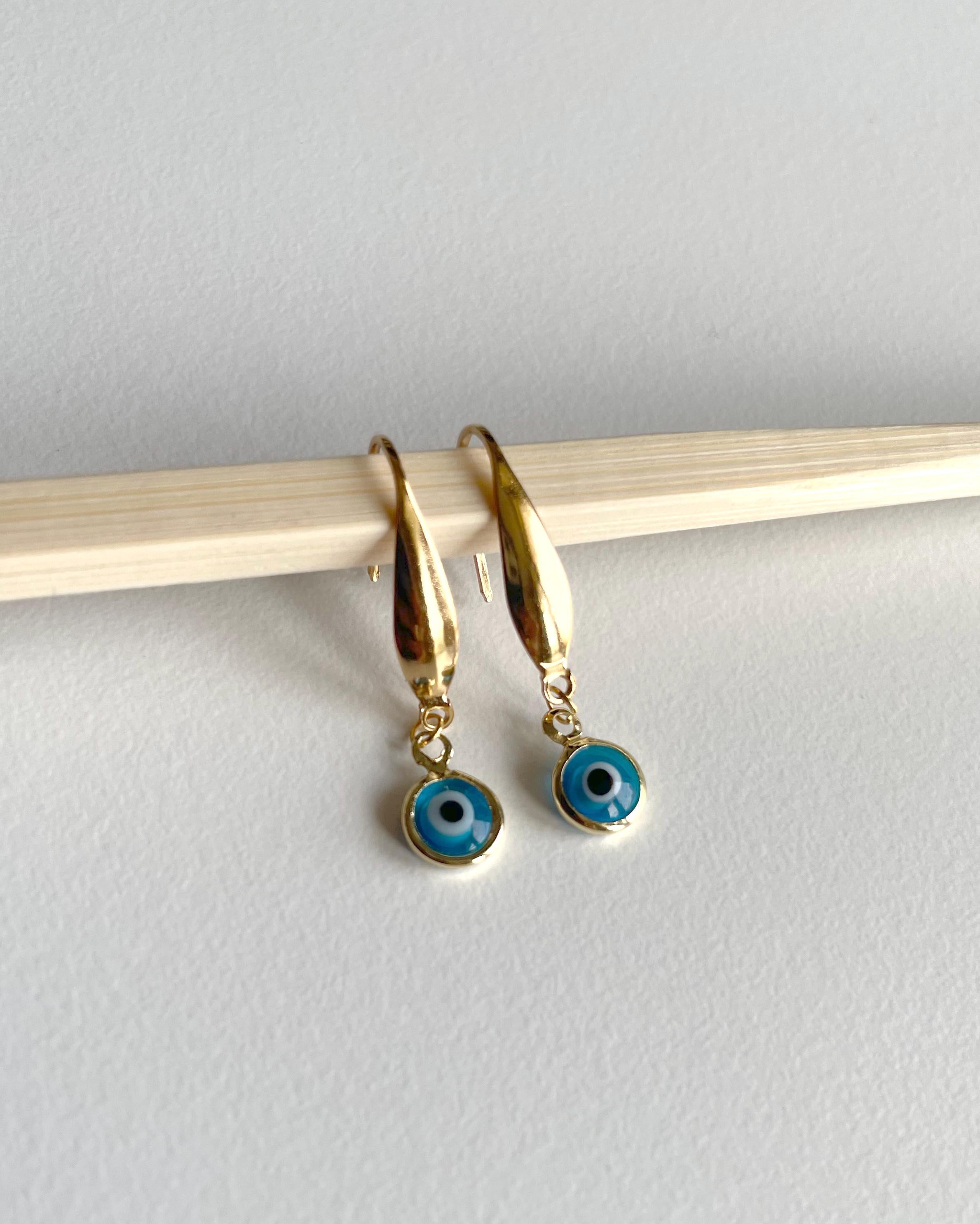 A pair of gold-tone dangle earrings with blue and white evil eye charms, hanging from a light-colored wooden stick against a neutral light gray background.
