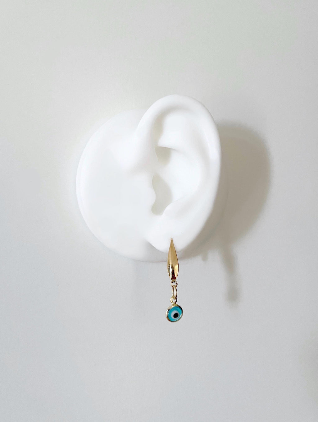 A gold-toned dangle earring with a small blue and black evil eye charm, displayed on a white ear-shaped stand against a light gray background.