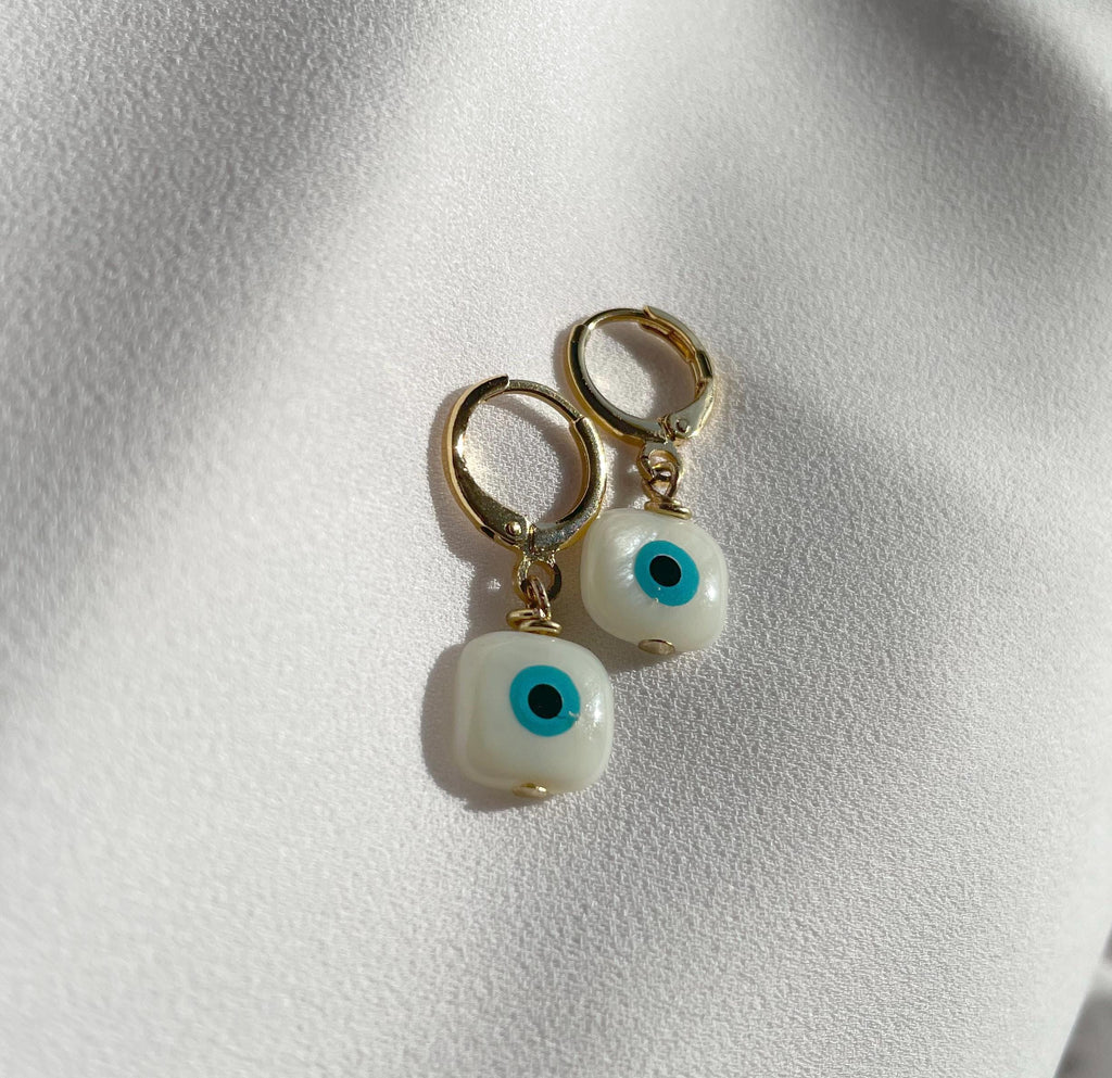 A pair of gold hoop earrings with white evil eye bead charms, displayed on a textured white fabric background.