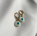 A pair of gold hoop earrings with white evil eye bead charms, displayed on a textured white fabric background.