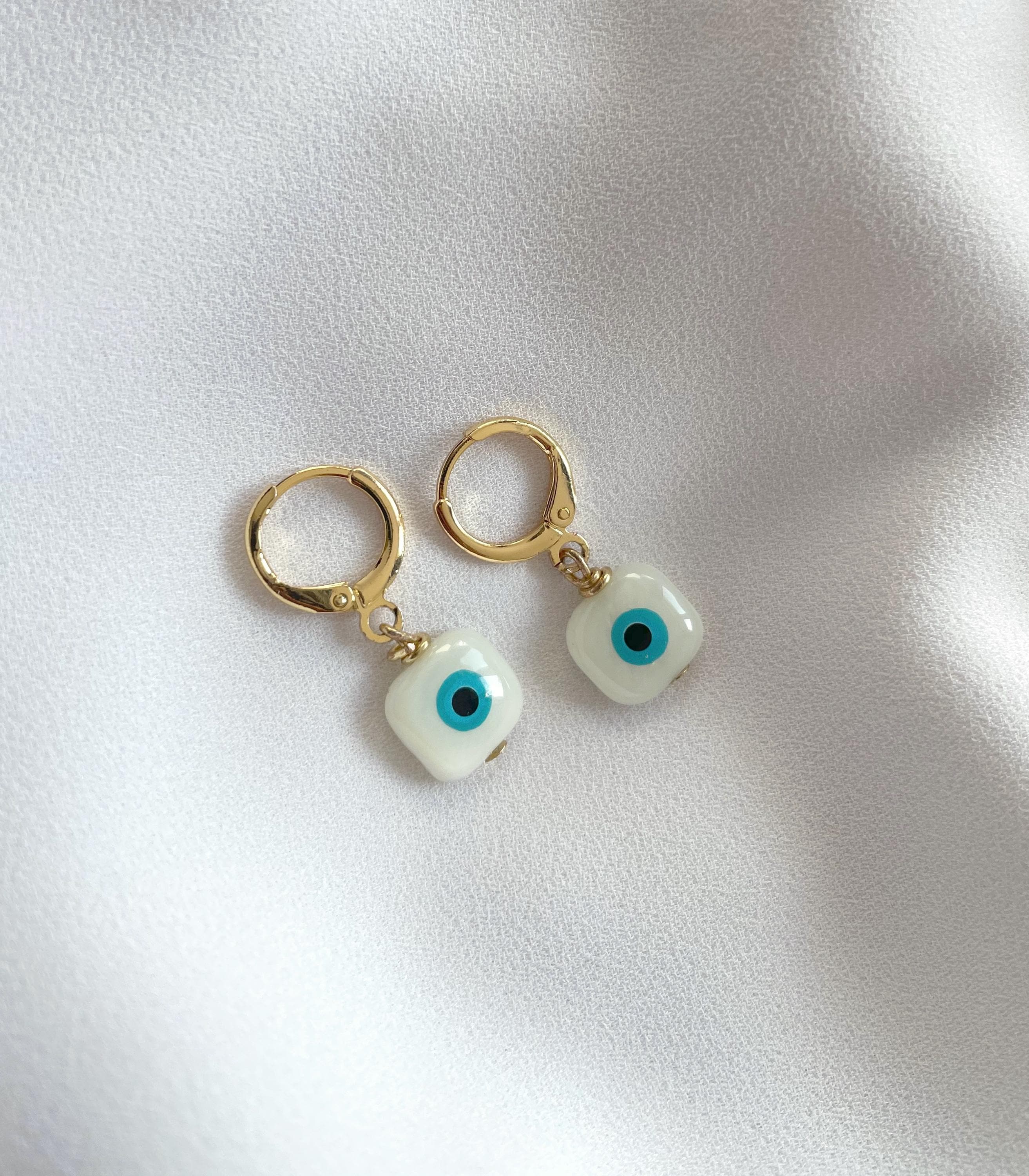 A pair of gold hoop earrings with small white evil eye charms, displayed on a soft, textured white fabric background.