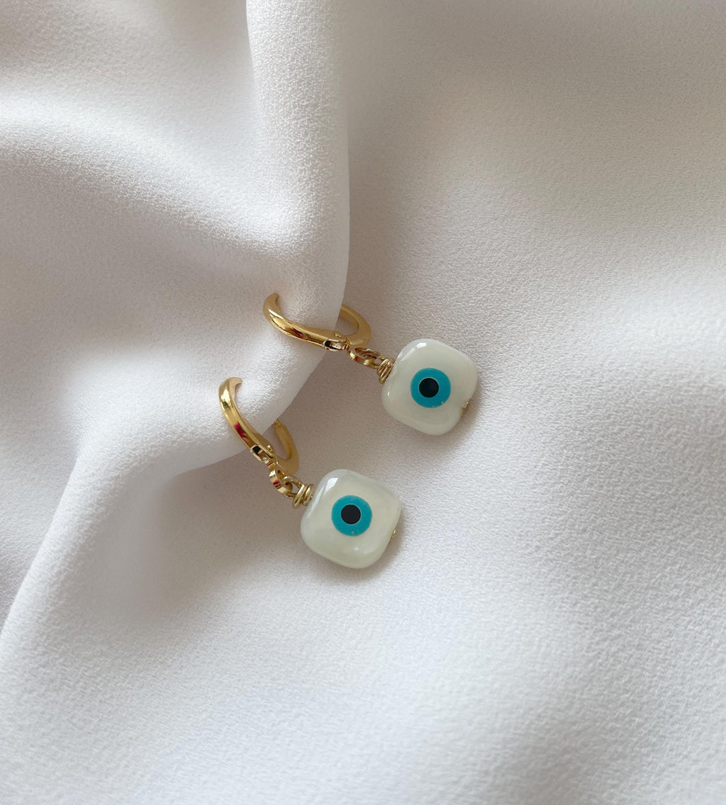 A pair of gold huggie hoop earrings with square white evil eye charms, displayed on a draped white fabric background.