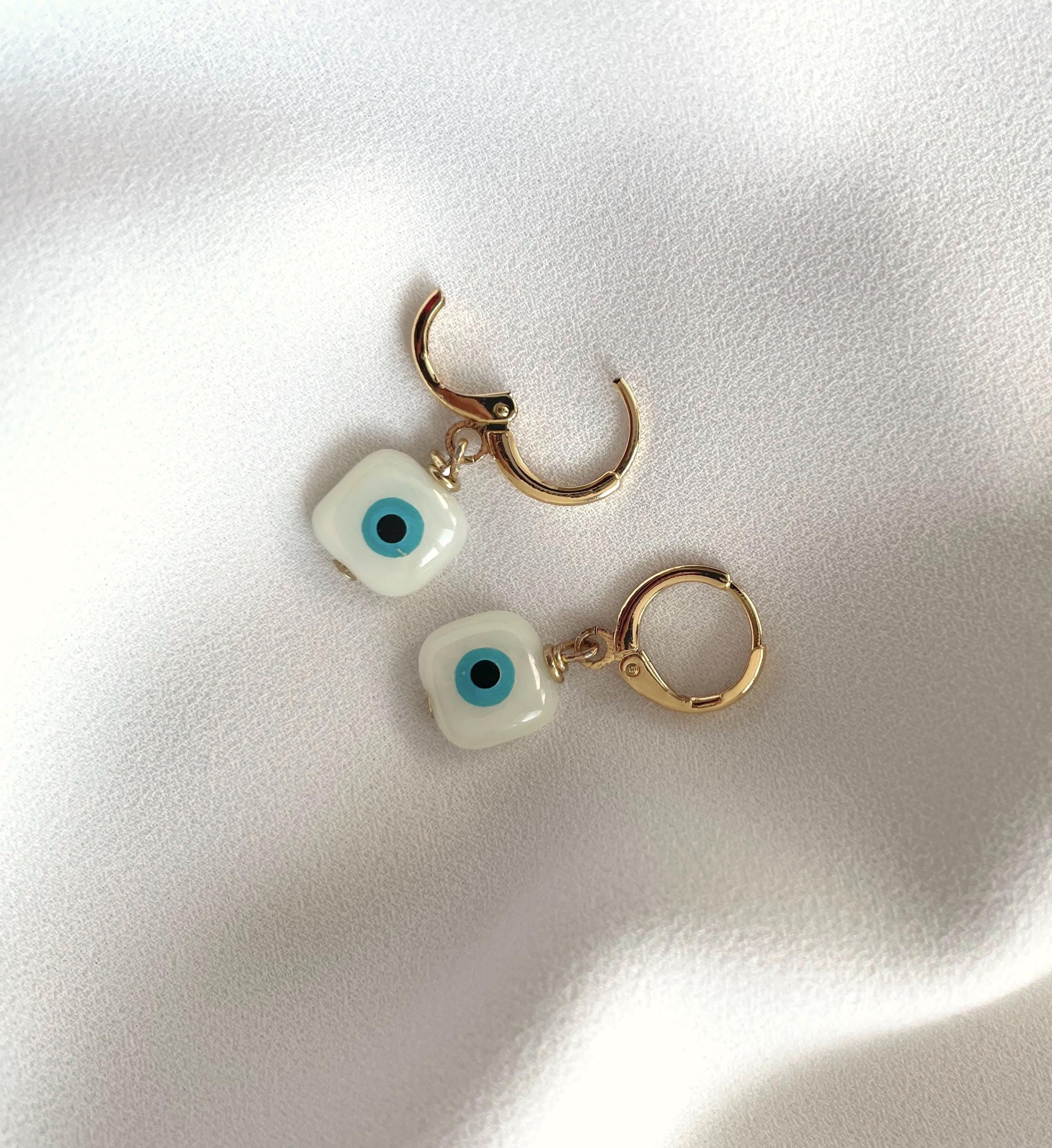 A pair of gold-tone huggie hoop earrings with dangling white evil eye charms featuring blue centers, displayed on a textured white fabric background.