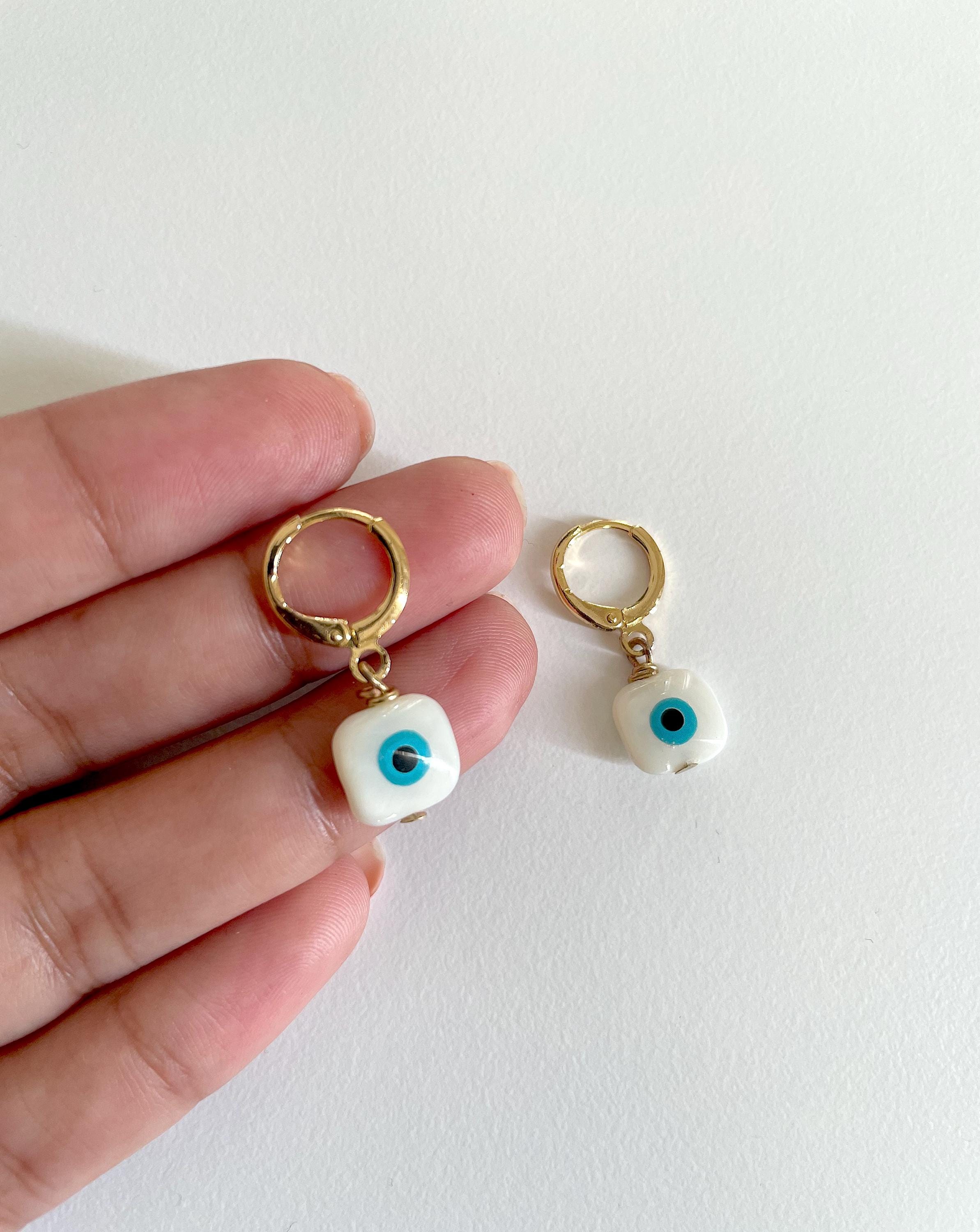 A close-up of a hand holding a pair of small gold hoop earrings with white square charms featuring blue and black evil eye designs, against a plain white background.