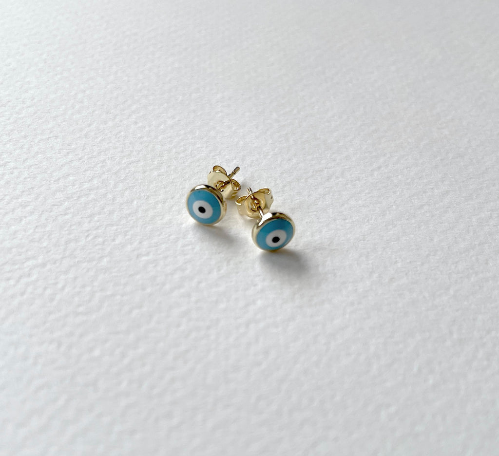 A pair of gold stud earrings with blue and white evil eye enamel details, resting on a textured white surface.