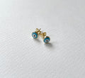 A pair of gold stud earrings with blue and white evil eye enamel details, resting on a textured white surface.