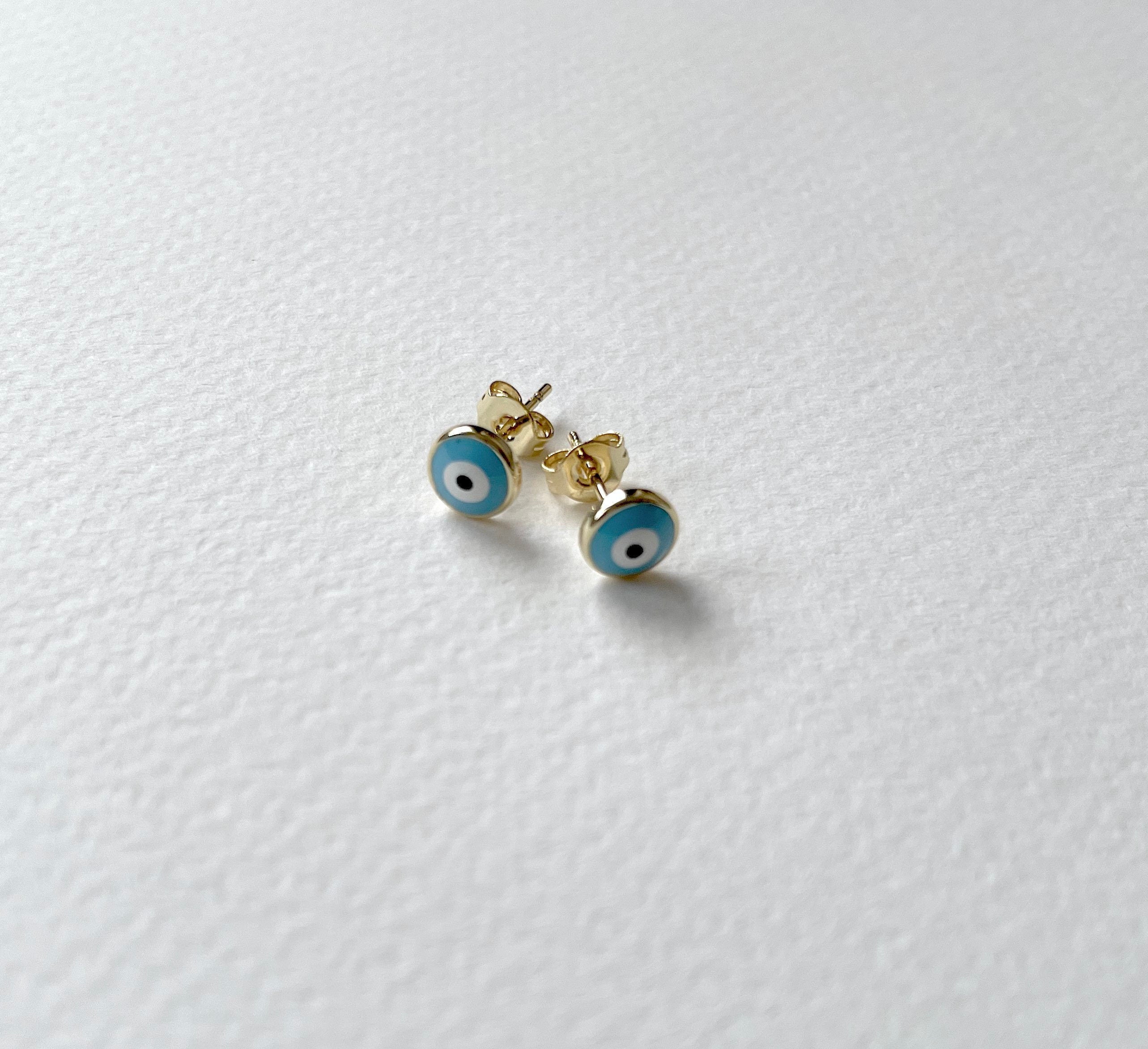 A pair of gold stud earrings with blue and white evil eye enamel details, resting on a textured white surface.