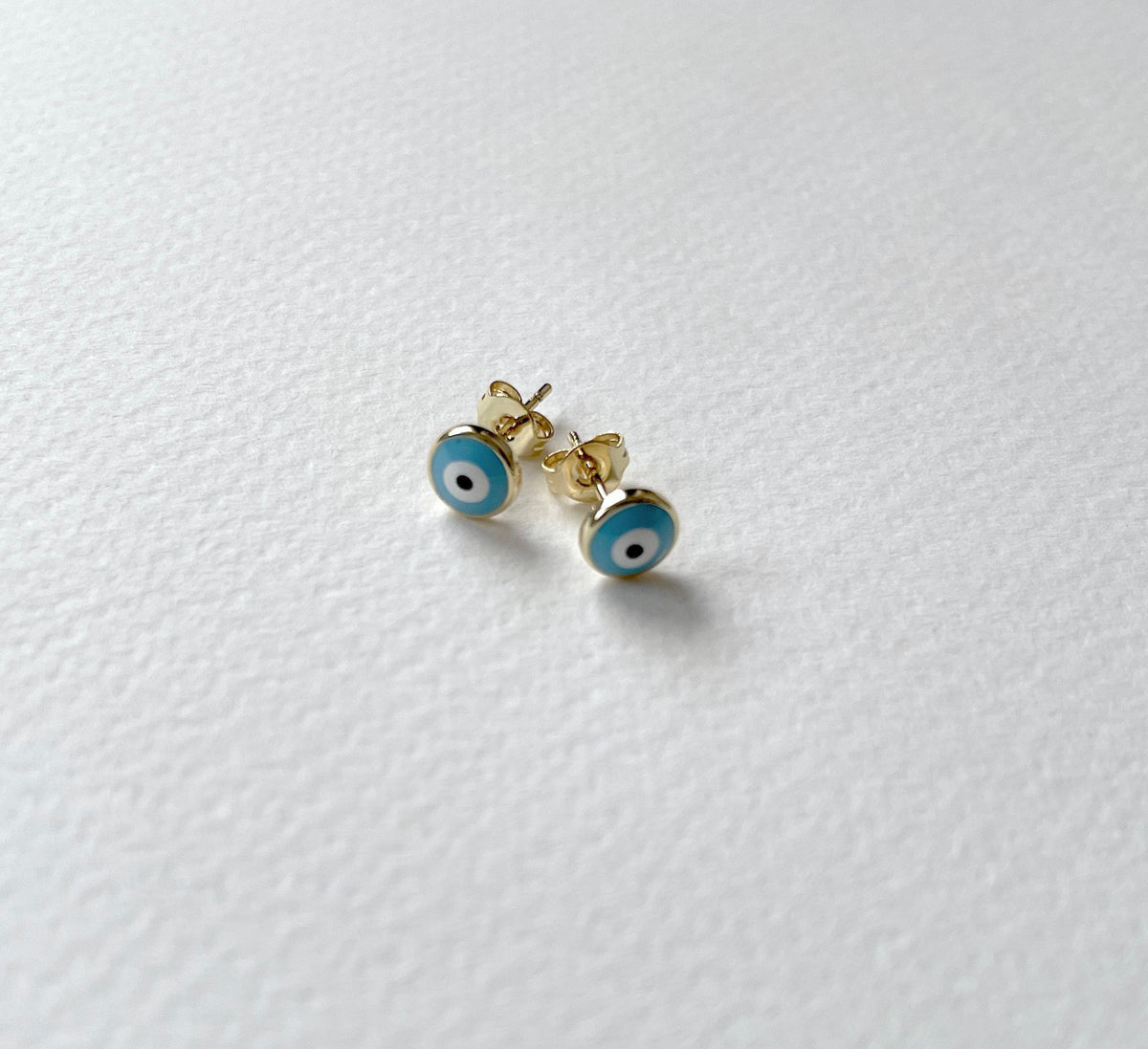 A pair of gold stud earrings with blue and white evil eye enamel details, resting on a textured white surface.