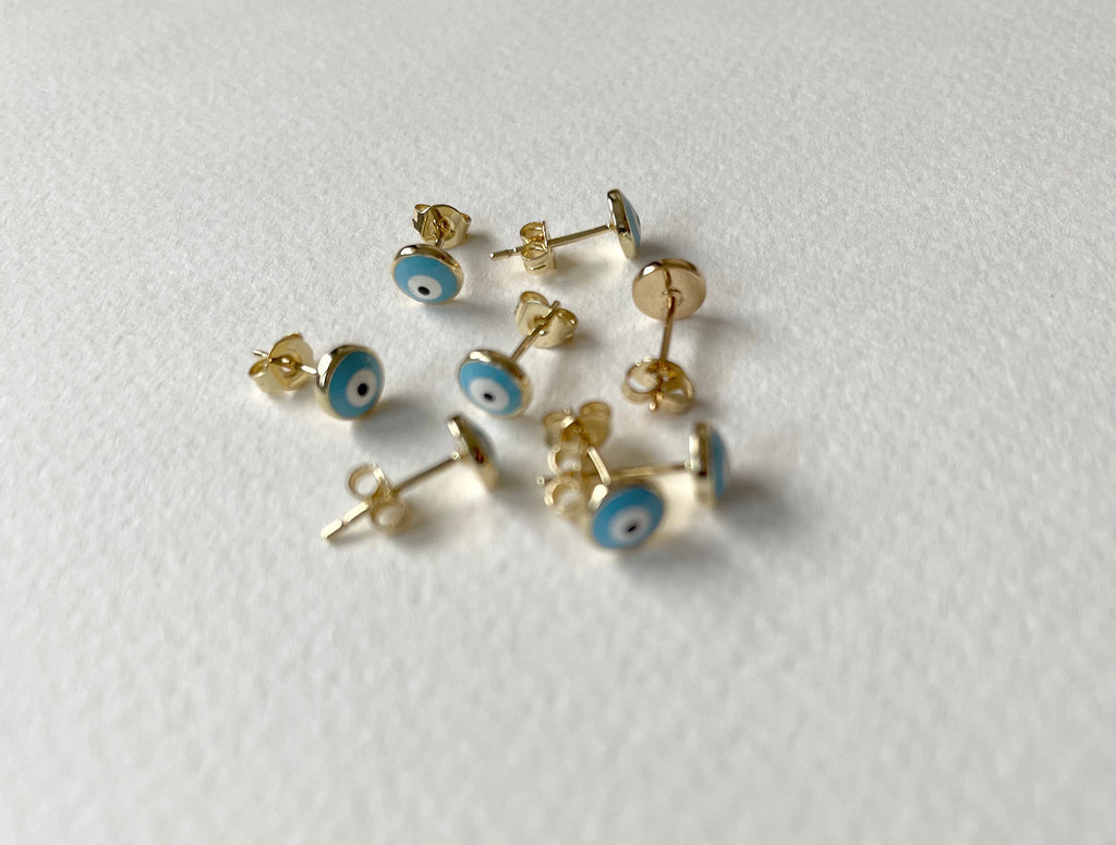 Several small gold stud earrings with blue and white evil eye motifs are scattered on a plain white textured surface.