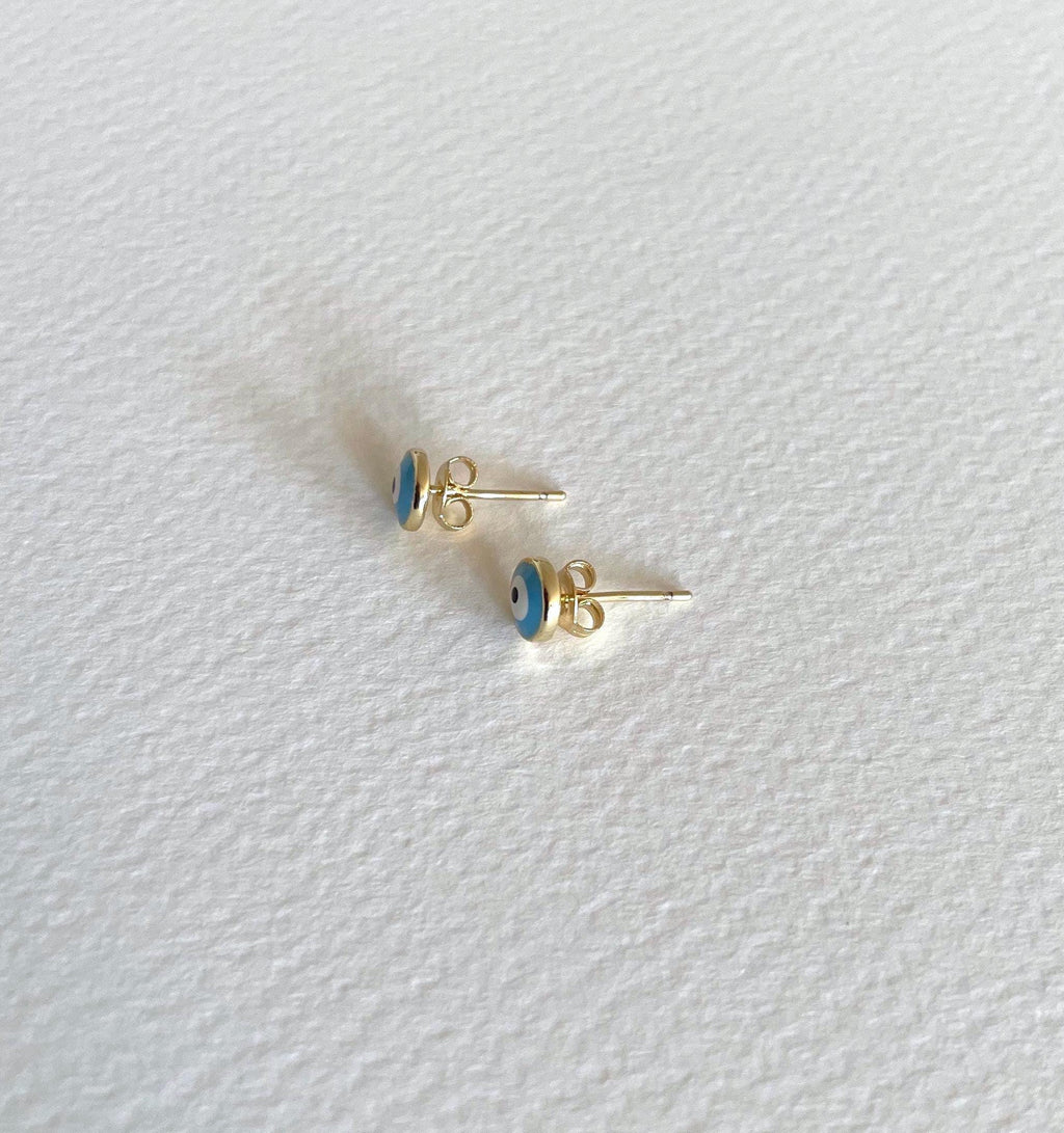 A pair of small gold stud earrings with blue and white eye motifs, displayed on a textured white surface.