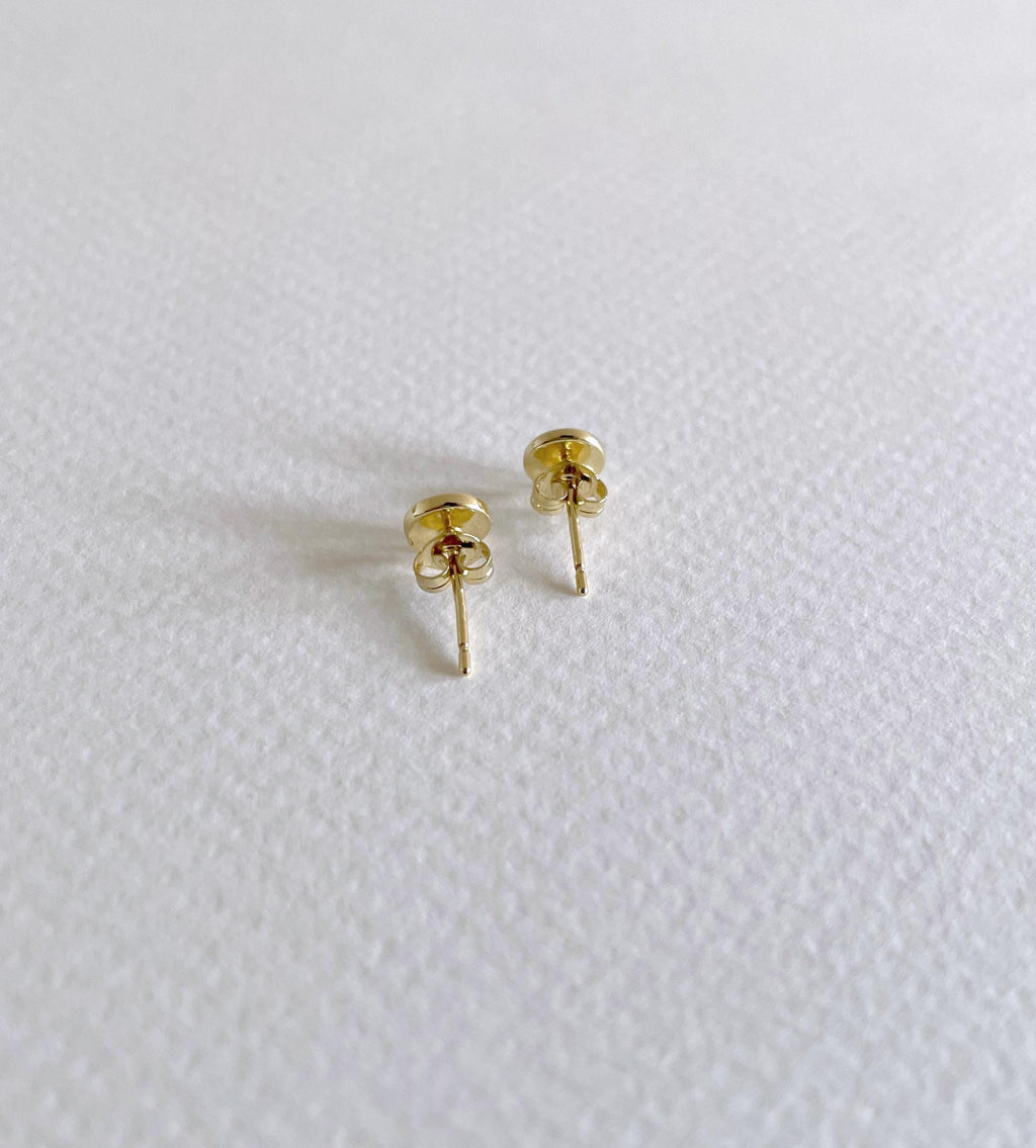 A pair of small gold evil eye stud earrings with butterfly backings, resting on a textured white surface.