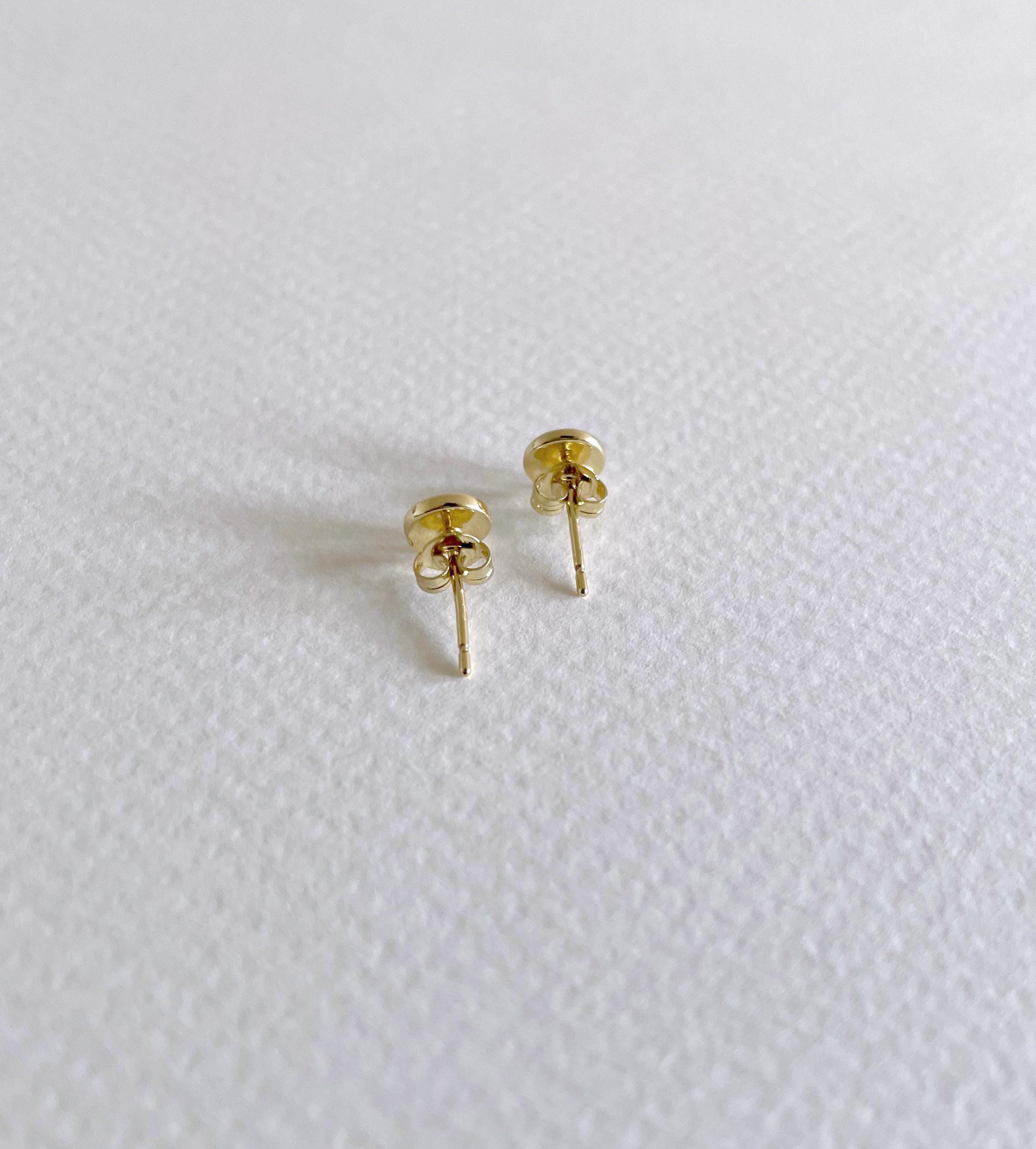 A pair of small gold evil eye stud earrings with butterfly backings, resting on a textured white surface.